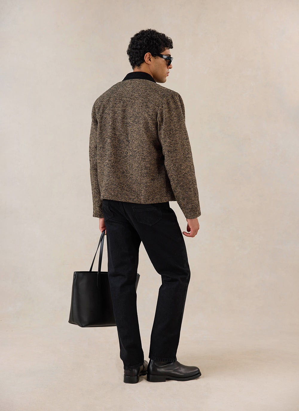 Rear view of a model wearing the Percival brown herringbone wool cropped jacket with black trousers and holding a tote bag.