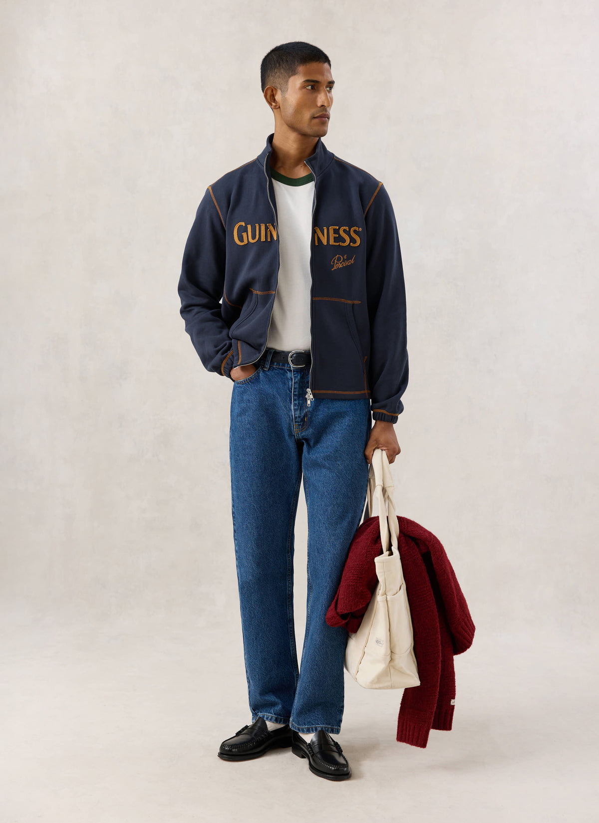 Model wearing the navy Guinness x Percival funnel neck sweatshirt, blue jeans, and holding a tote bag.