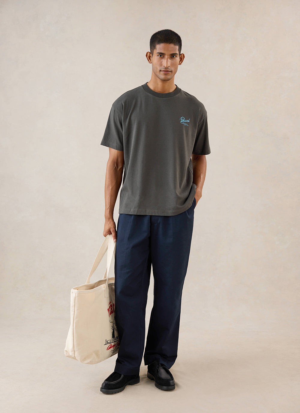 A model wears the charcoal Percival Dawson boxy t-shirt, styled with navy trousers and holding a tote bag.