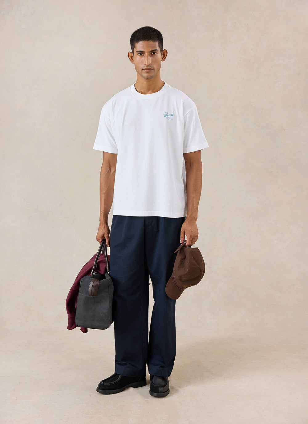 A model wears the white Percival Dawson t-shirt, styled with dark trousers, a holdall and a cap in a studio.