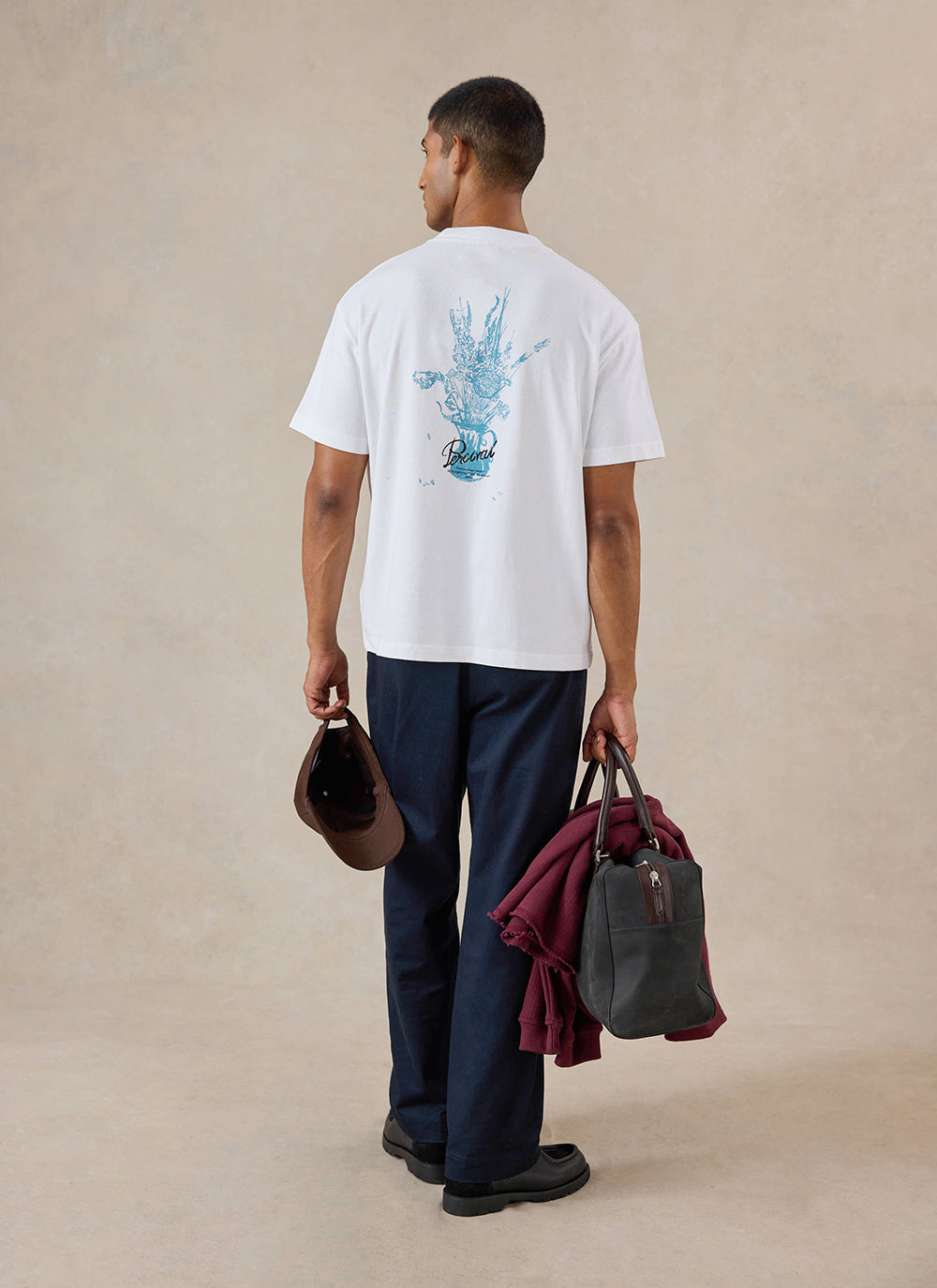 A model from behind wears the white Percival Dawson T-Shirt with blue back graphics, styled with dark trousers and a bag.
