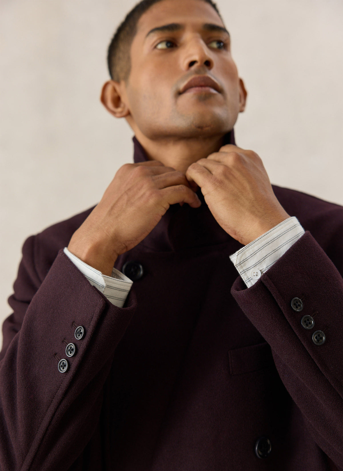 A close-up of a model adjusting the collar of the Percival dark brown overcoat over a striped shirt.