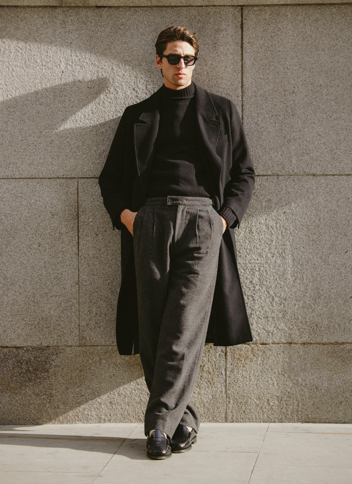 A model wearing a black Percival double-breasted overcoat, black roll-neck jumper, and charcoal tailored trousers outdoors.