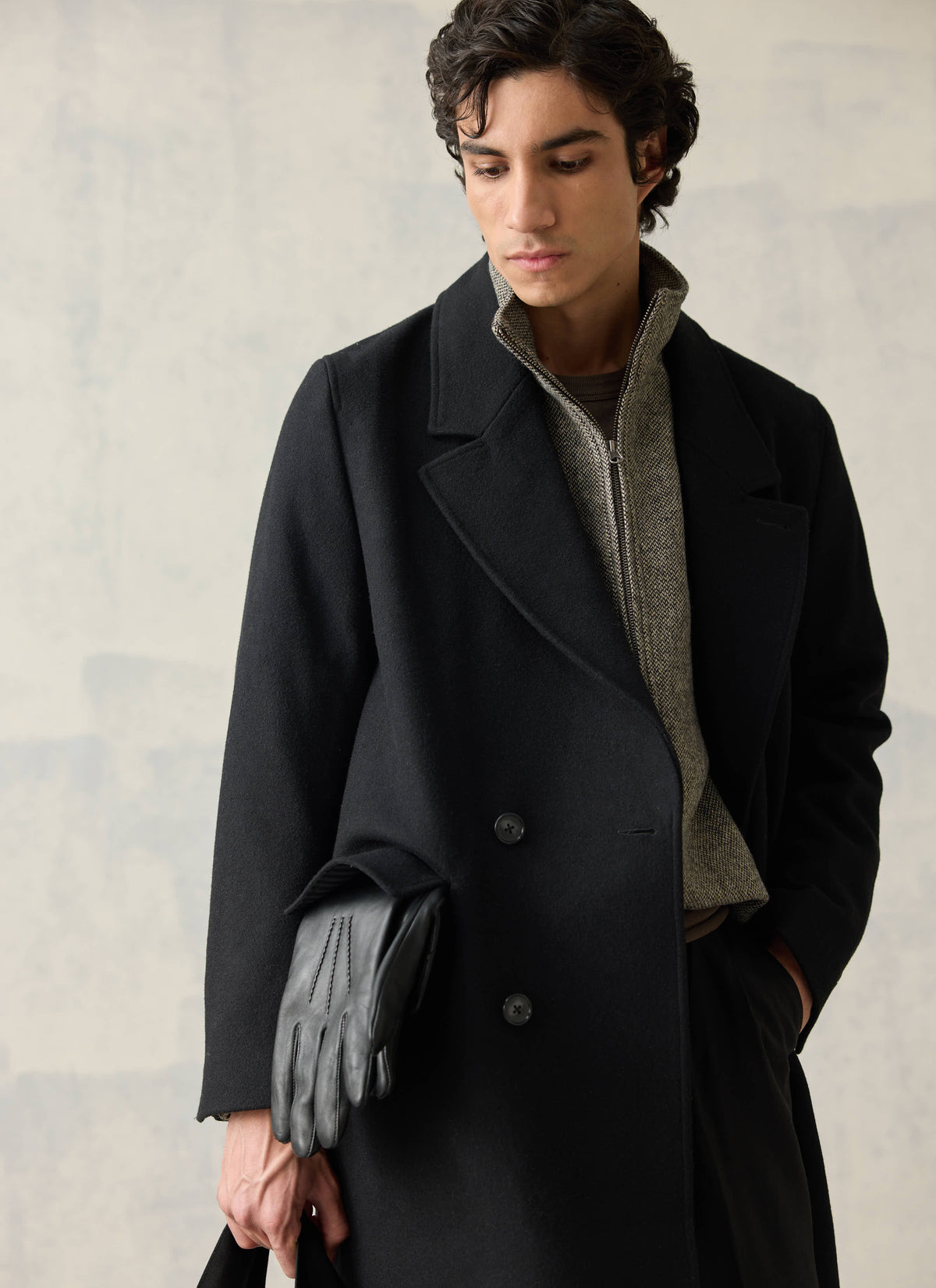A model wearing a black wool blend double-breasted overcoat over a patterned zip jumper and holding black leather gloves.