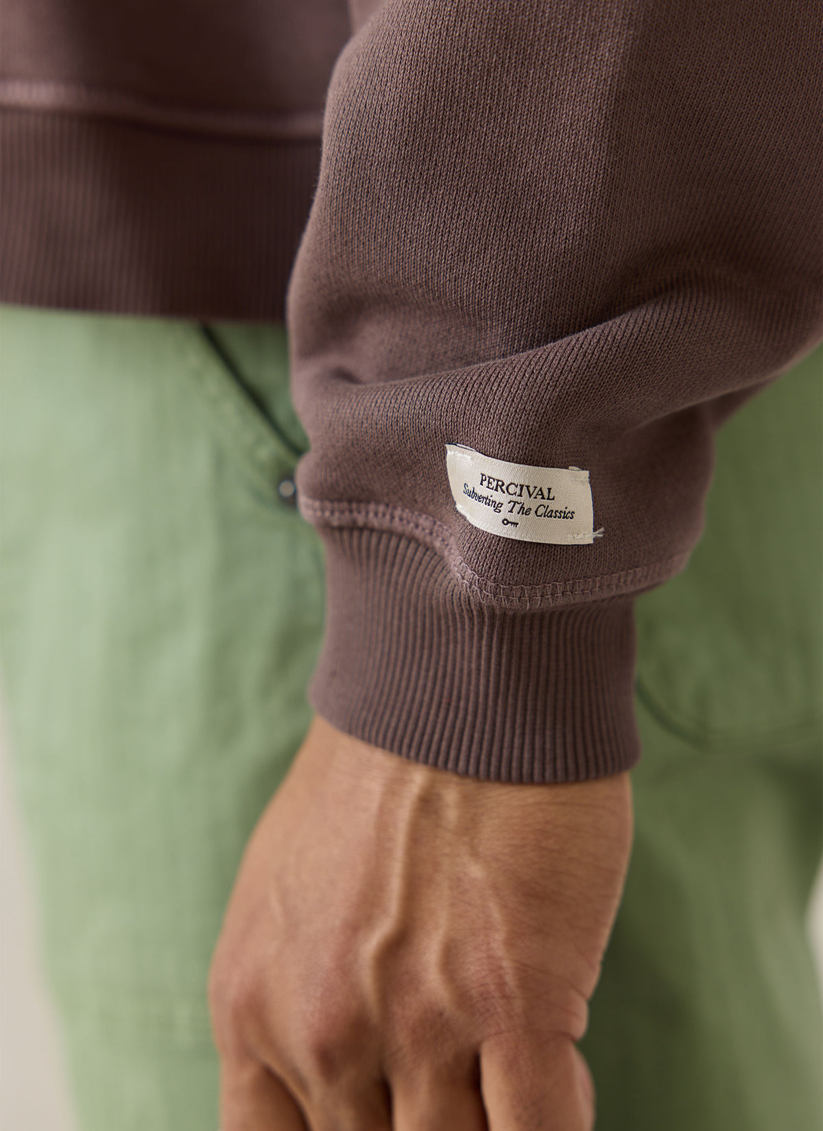 A close-up of the brown Percival Dudley sweatshirt sleeve showing the ribbed cuff and branded logo label.