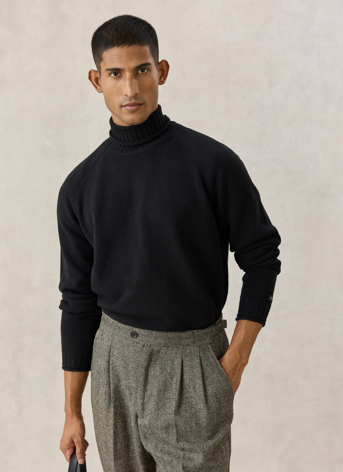A model wearing the Percival black wool roll-neck jumper with raglan sleeves, paired with patterned grey trousers.