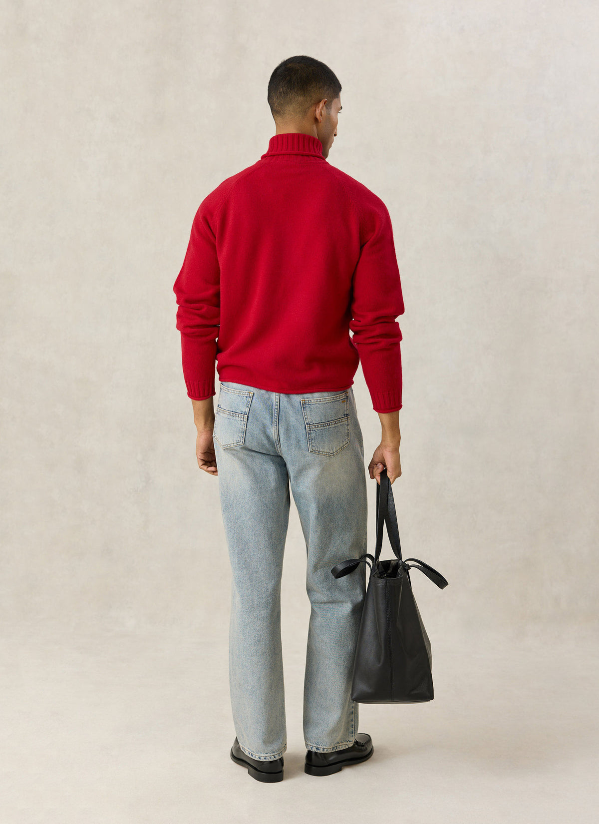A model from the back wearing the red Percival wool roll-neck jumper, light wash jeans, and holding a black leather tote bag.