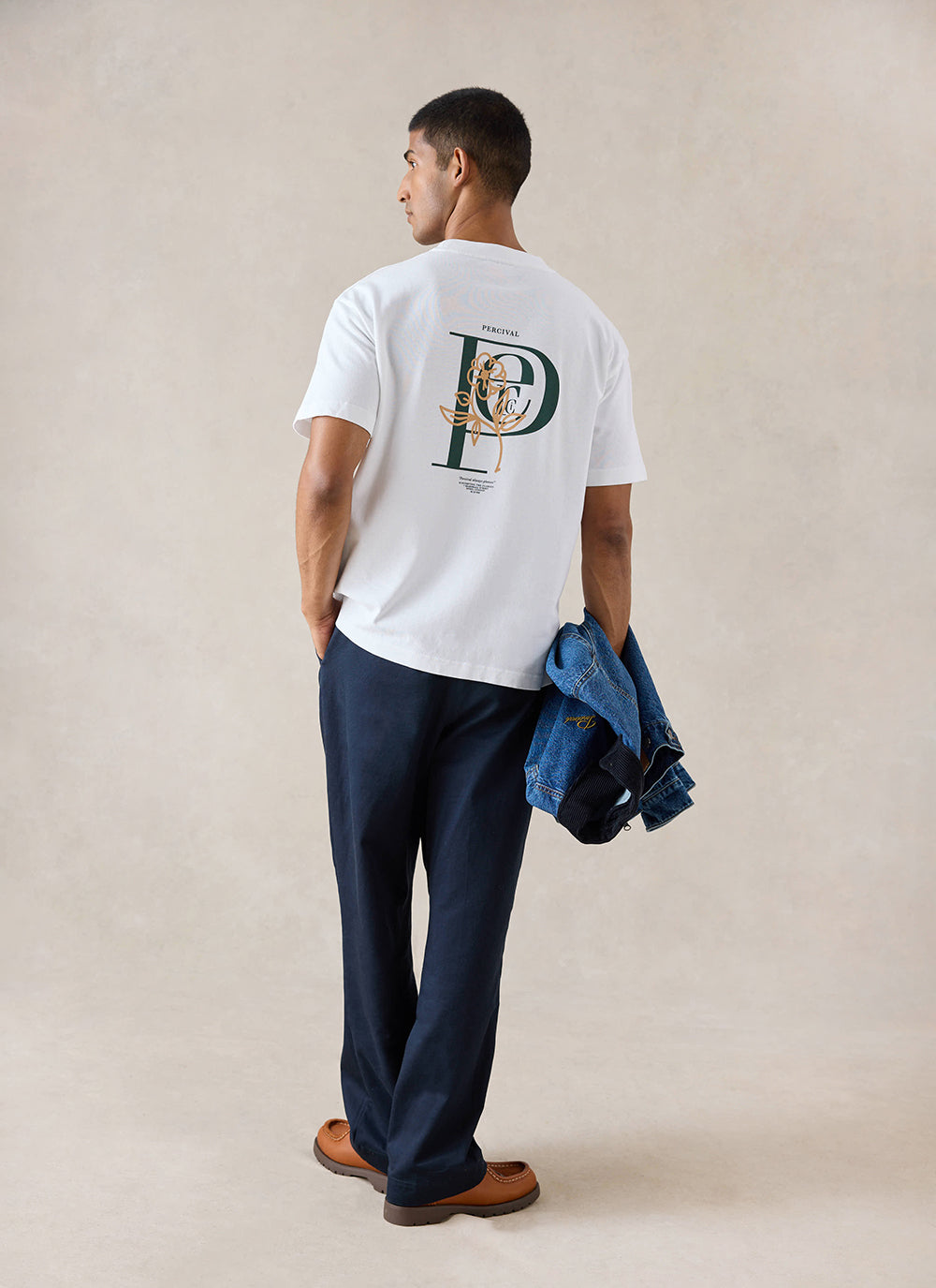 A model wears the white Percival Earl T-Shirt, showing the back graphic print, styled with navy trousers.