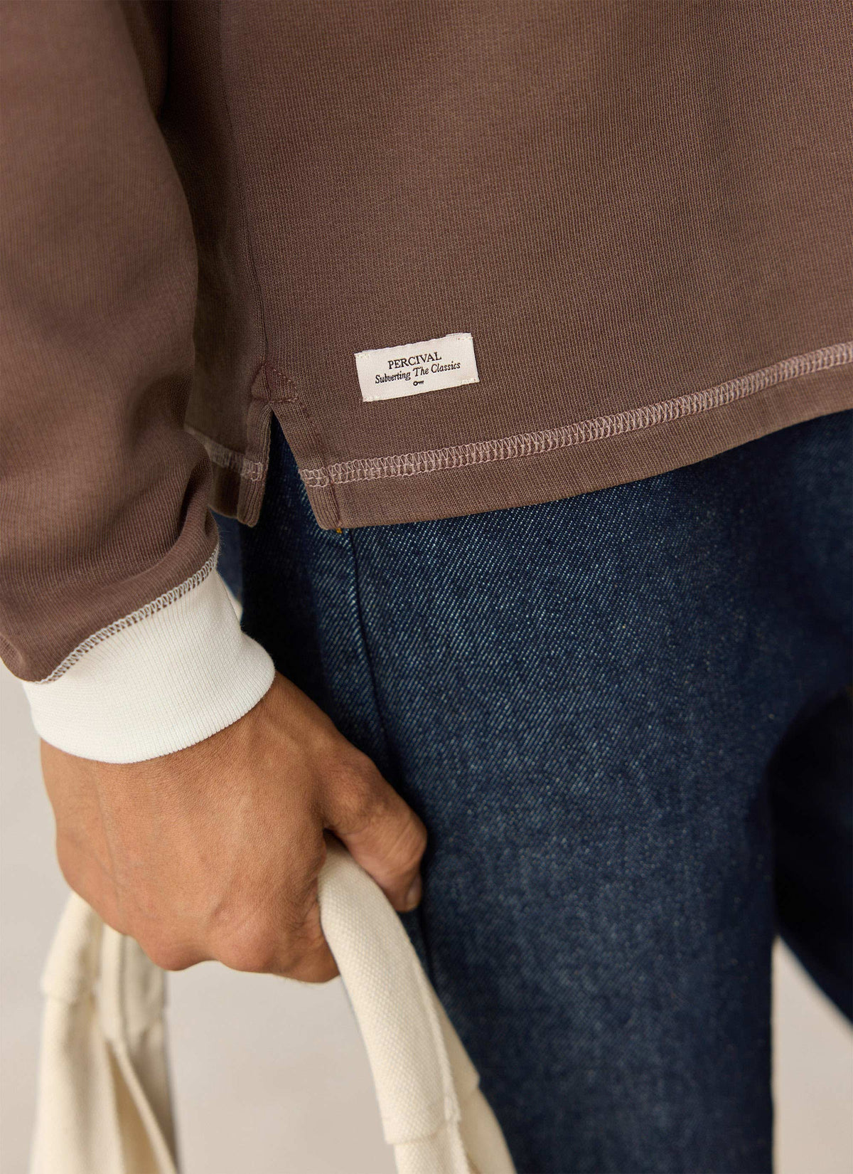 Close-up of the brown Percival cotton twill rugby shirt showing the hem label, white cuff, and denim jeans.