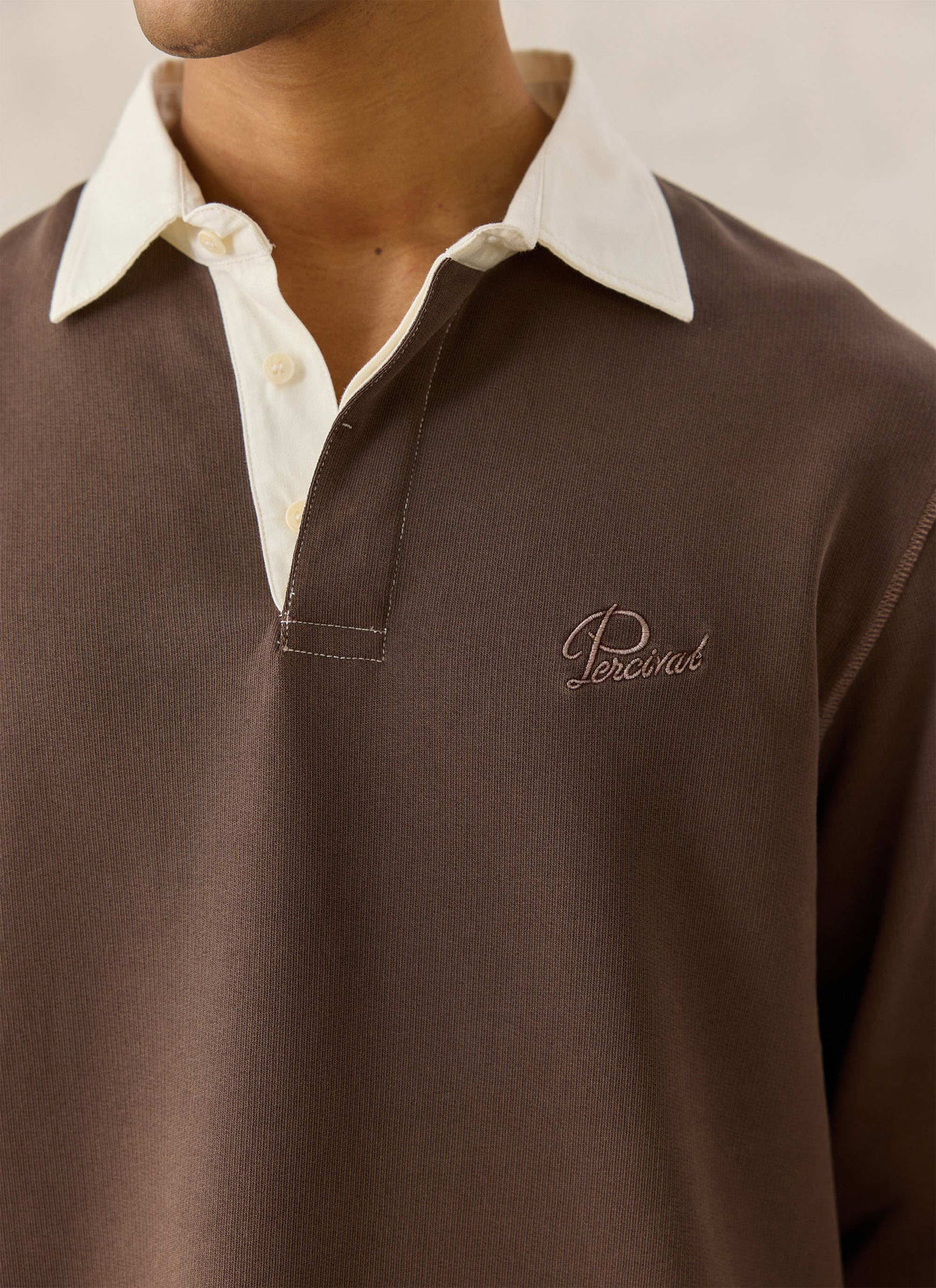 A close-up of the Percival brown cotton twill rugby polo shirt showing the contrasting cream collar and chest embroidery.