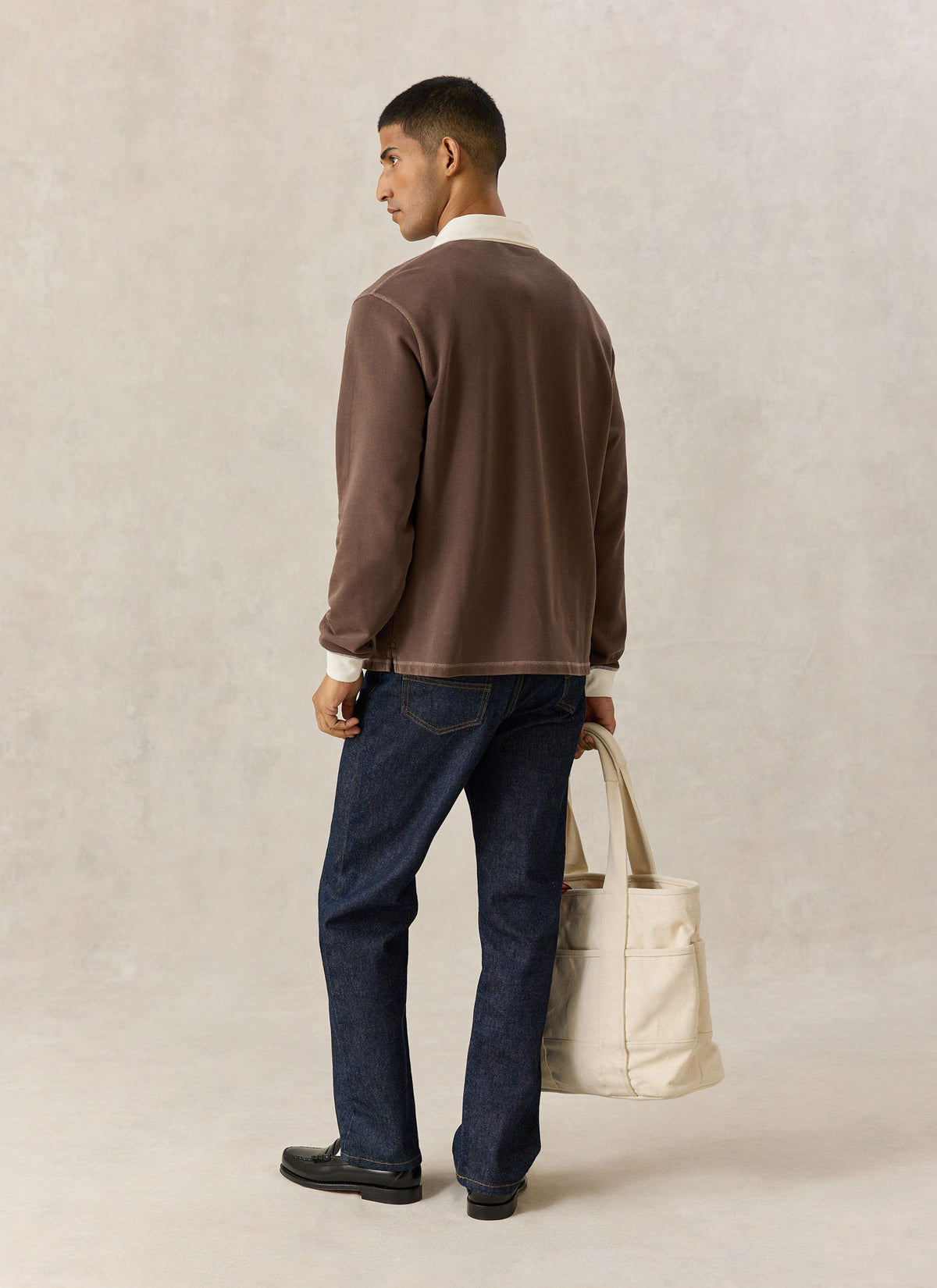 A model wearing the brown Percival cotton rugby polo shirt, dark jeans, and carrying a canvas tote bag.