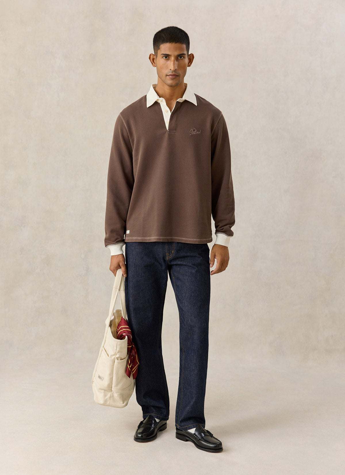A model wearing the brown Percival rugby polo shirt with cream collar, dark jeans and a tote bag.