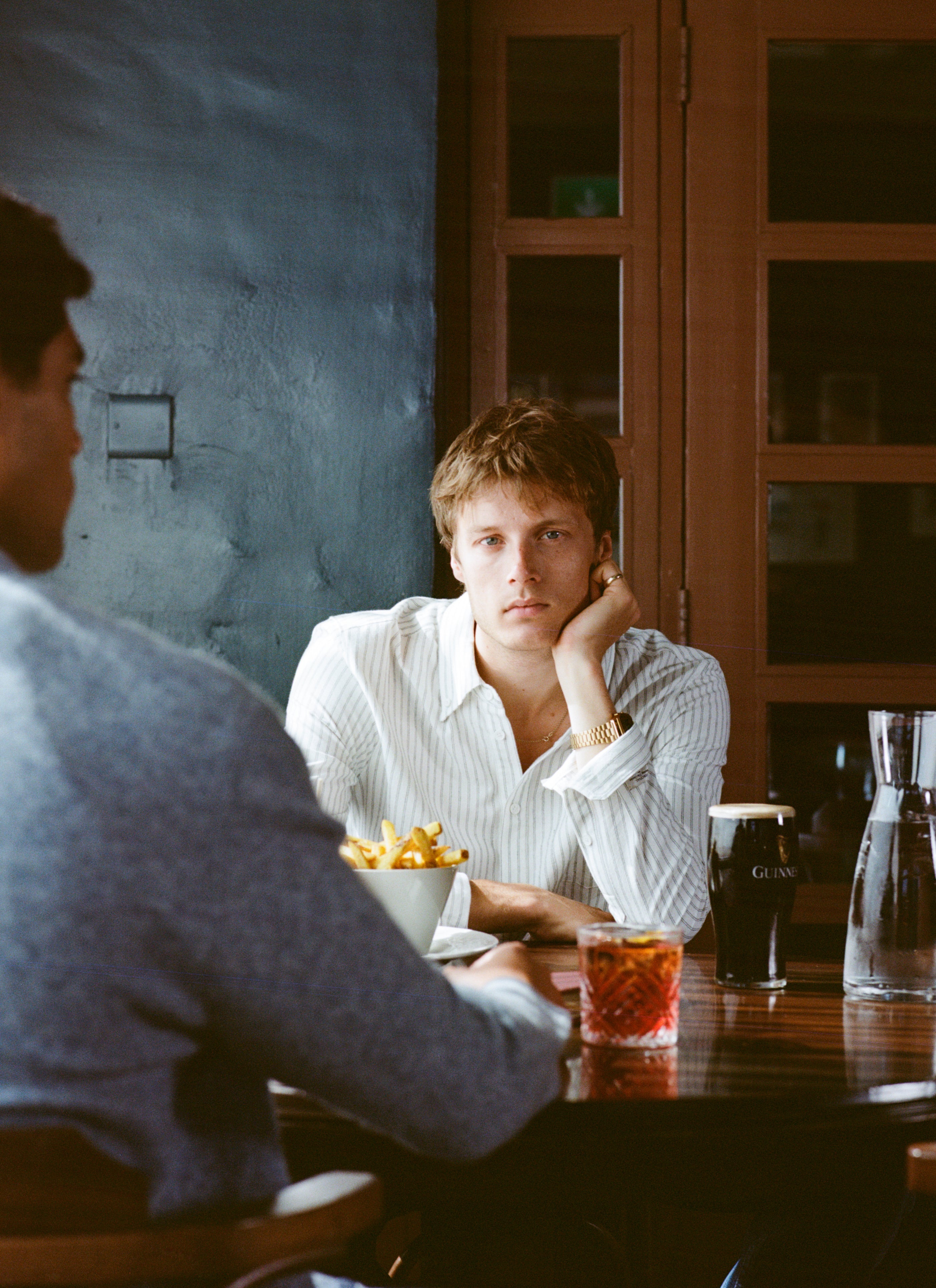 A model wears the white Percival Fabian Striped Oxford Shirt, seated at a pub table with a drink and chips.