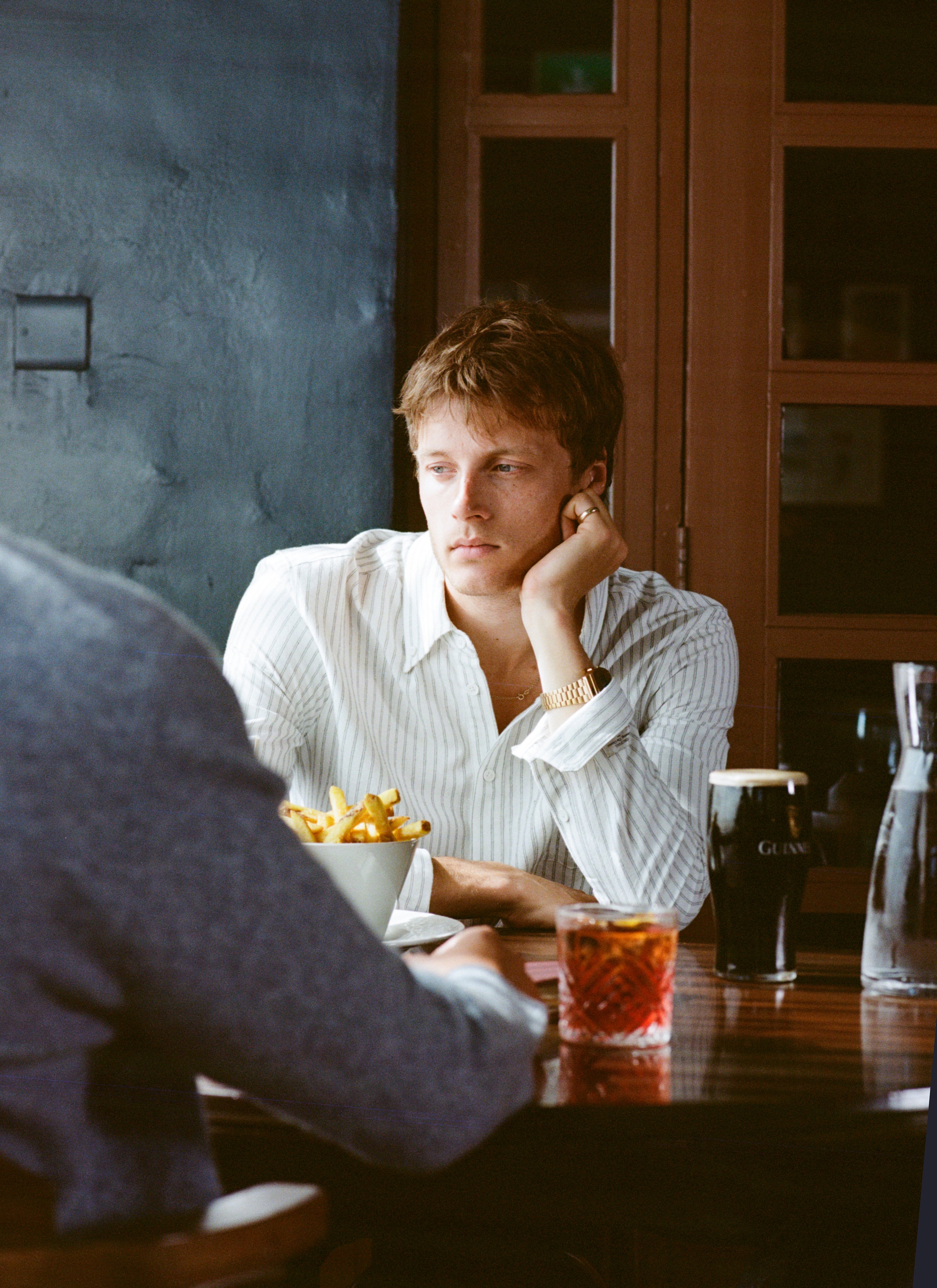 A man sits at a pub table wearing the white striped Fabian Oxford shirt from Percival, resting his head on his hand.