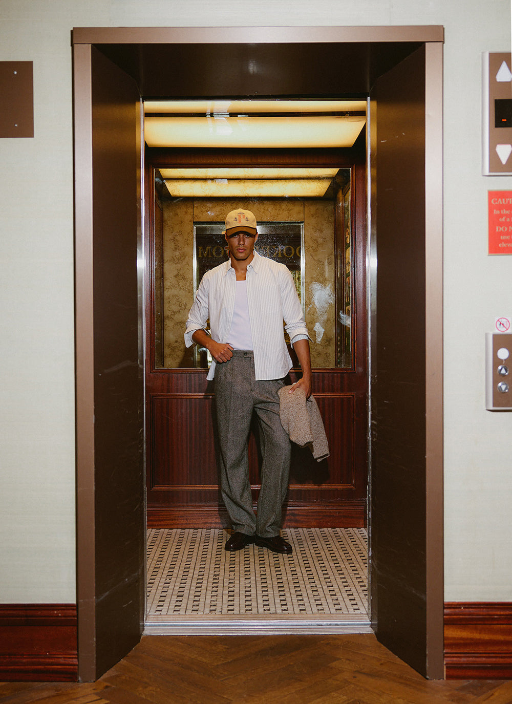 A model wears the white Percival striped Oxford shirt, styled inside an elevator with grey trousers and a cap.