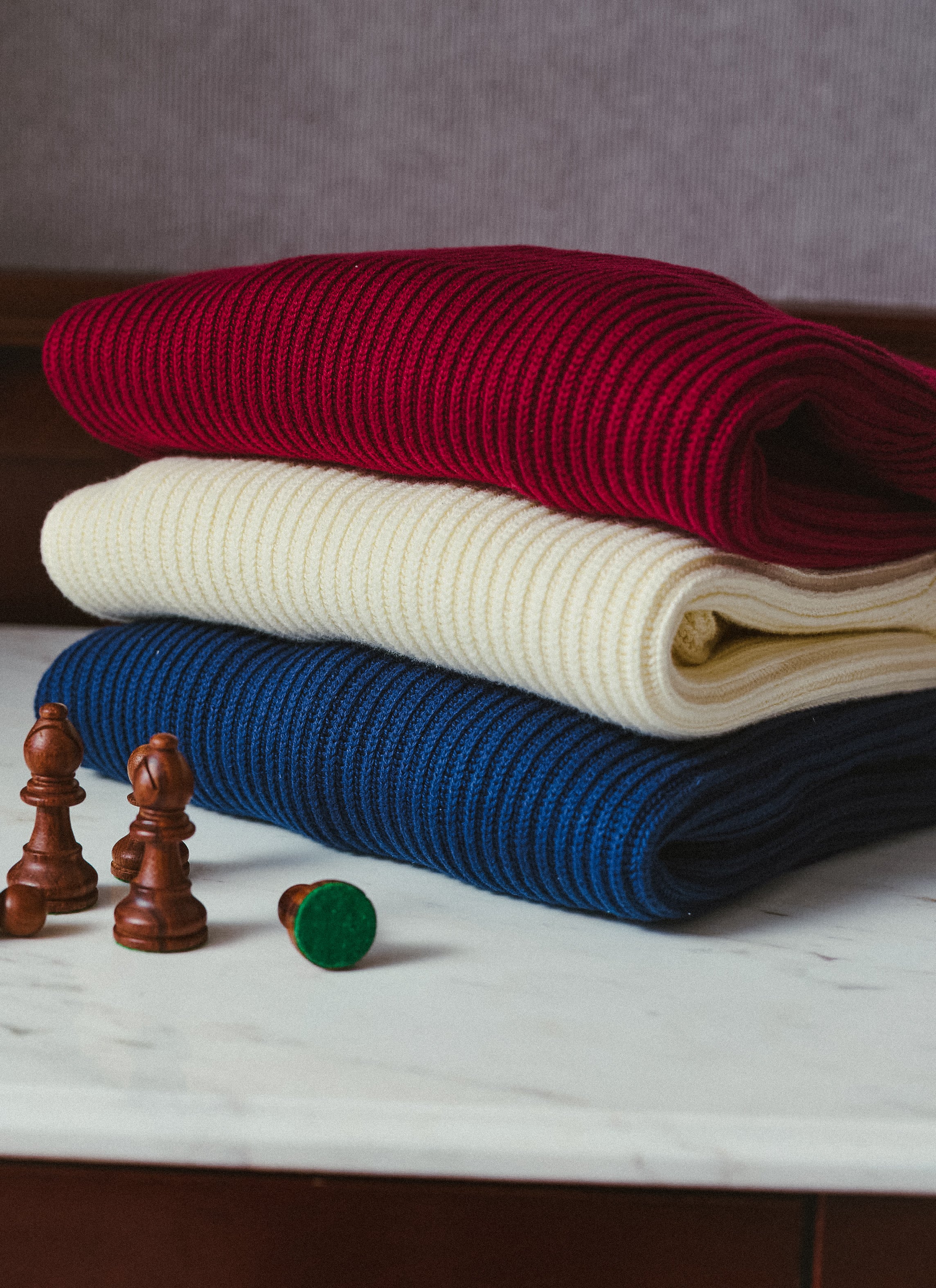 A stack of Percival fisherman jumpers in burgundy, cream and navy, displayed on a surface alongside wooden chess pieces.