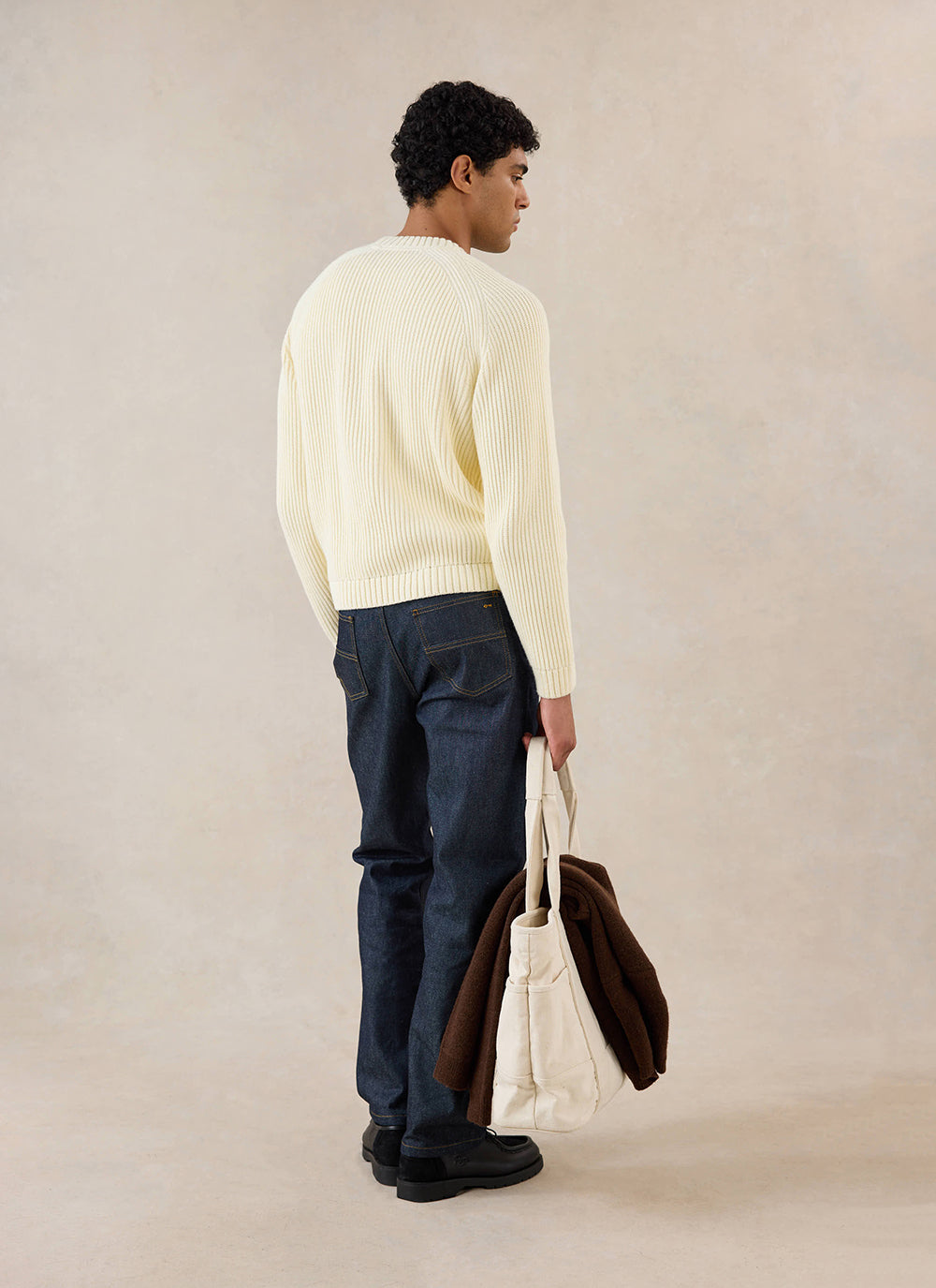 A model wears the cream Percival Fisherman Knitted Jumper from the back, styled with dark jeans and a canvas tote bag.