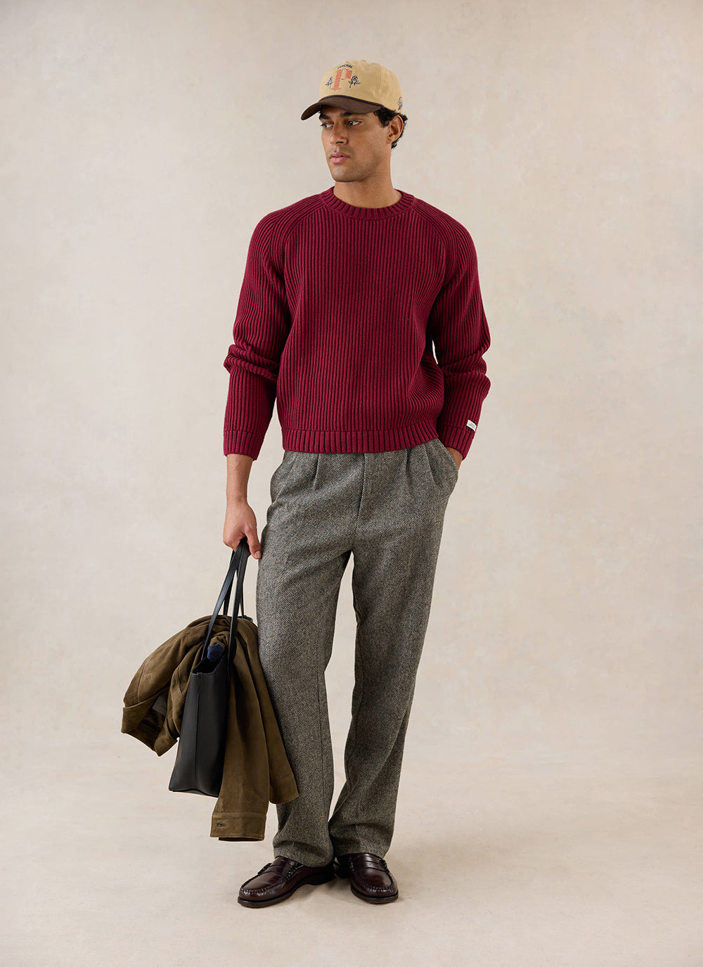 A model wears the burgundy Fisherman Raglan Jumper from Percival, styled with grey trousers, a cap, and a leather tote bag.