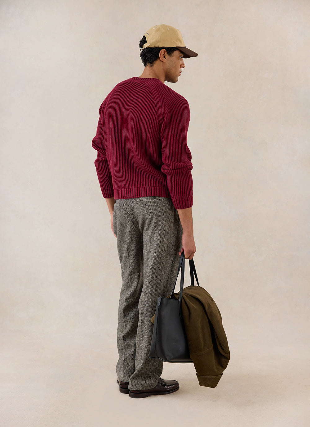 A rear view of a model wearing the merlot Fisherman Knitted Jumper from Percival, holding a black tote bag and a jacket.