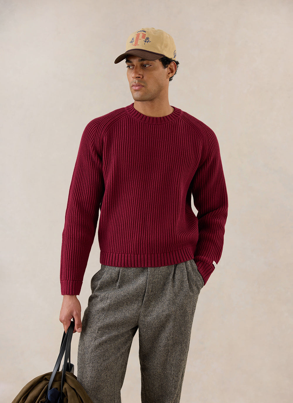 A model wears the burgundy Fisherman Raglan Jumper from Percival, styled with patterned trousers and a cap.