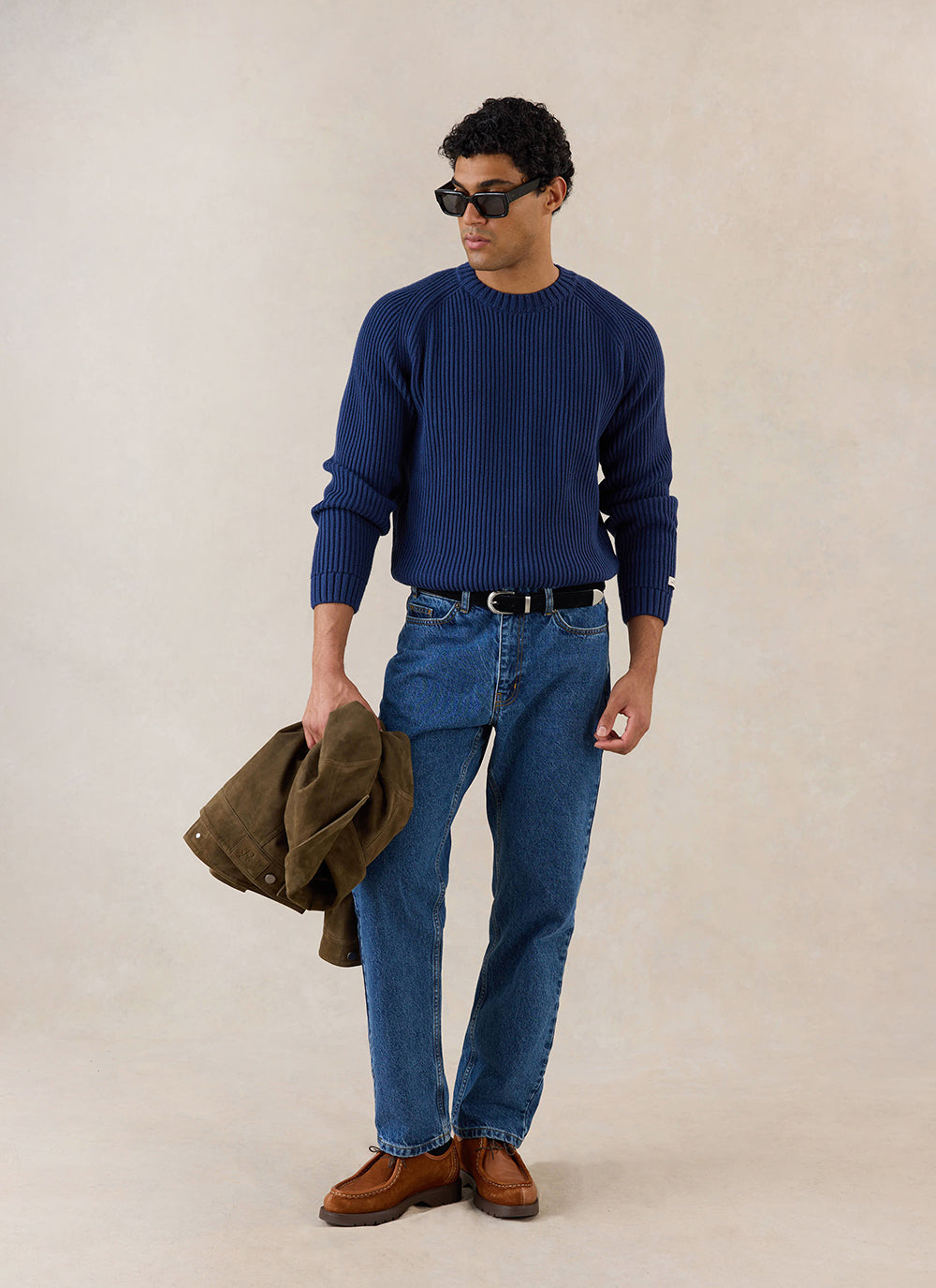 A model wears the navy Fisherman Knitted Jumper from Percival with blue jeans and brown shoes, holding a jacket.
