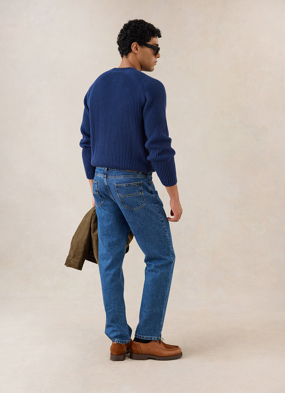 A model wears the navy Percival Fisherman Raglan Jumper from the back, styled with blue jeans and brown shoes.