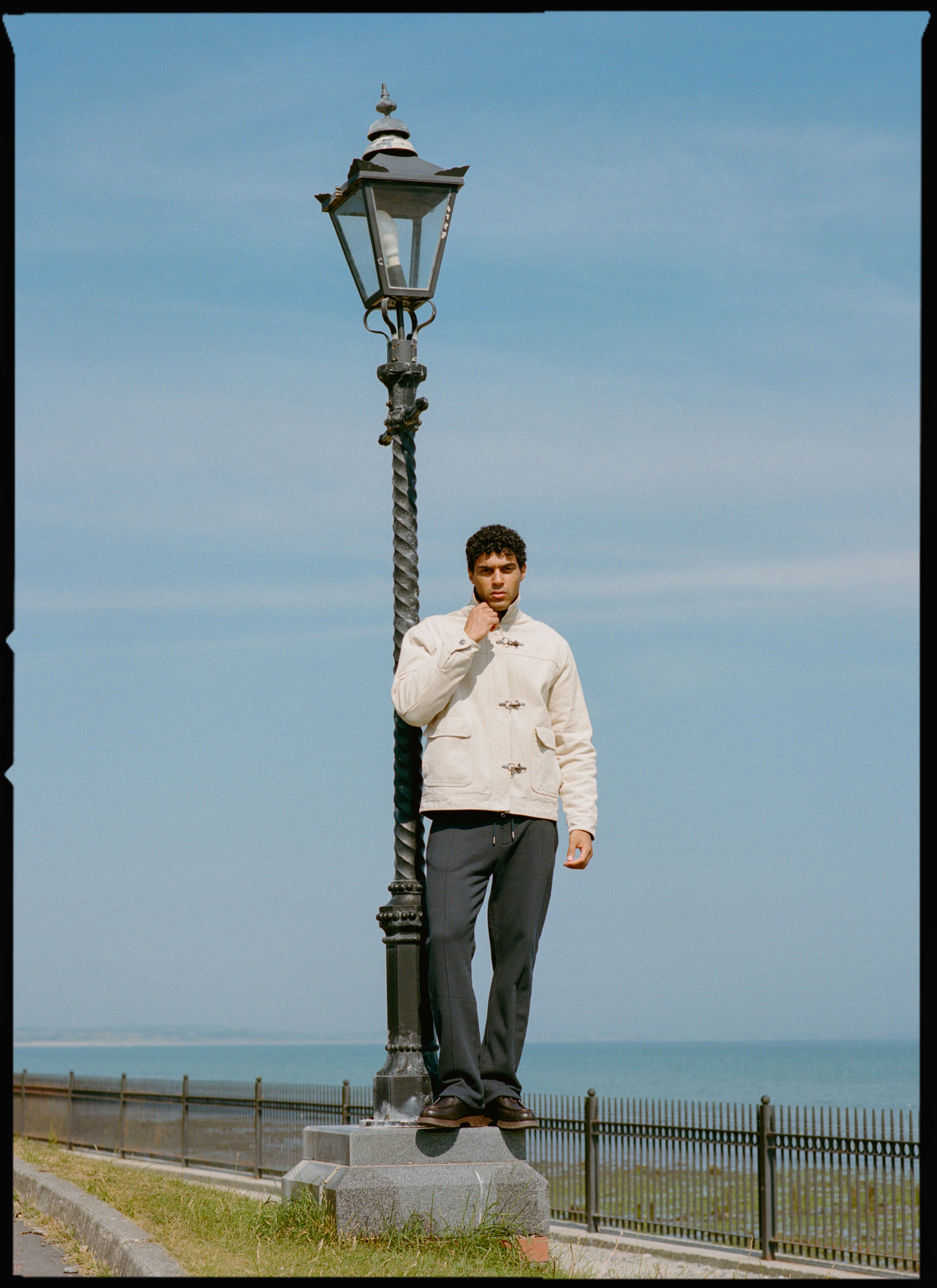 A model wears the cream Percival Fontane Fireman Clasp Jacket by a coastal lamppost.