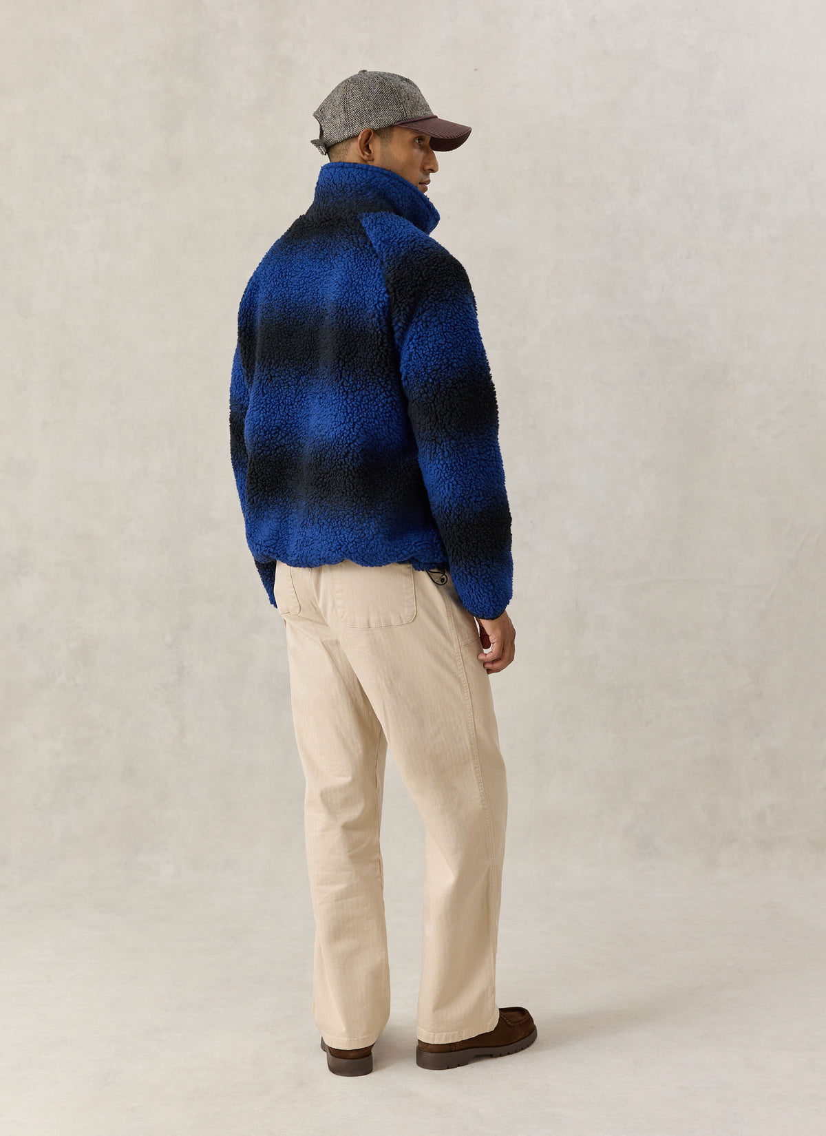 A model wearing the Percival ombre blue wool-blend fleece jacket from the back, paired with light trousers and a cap.