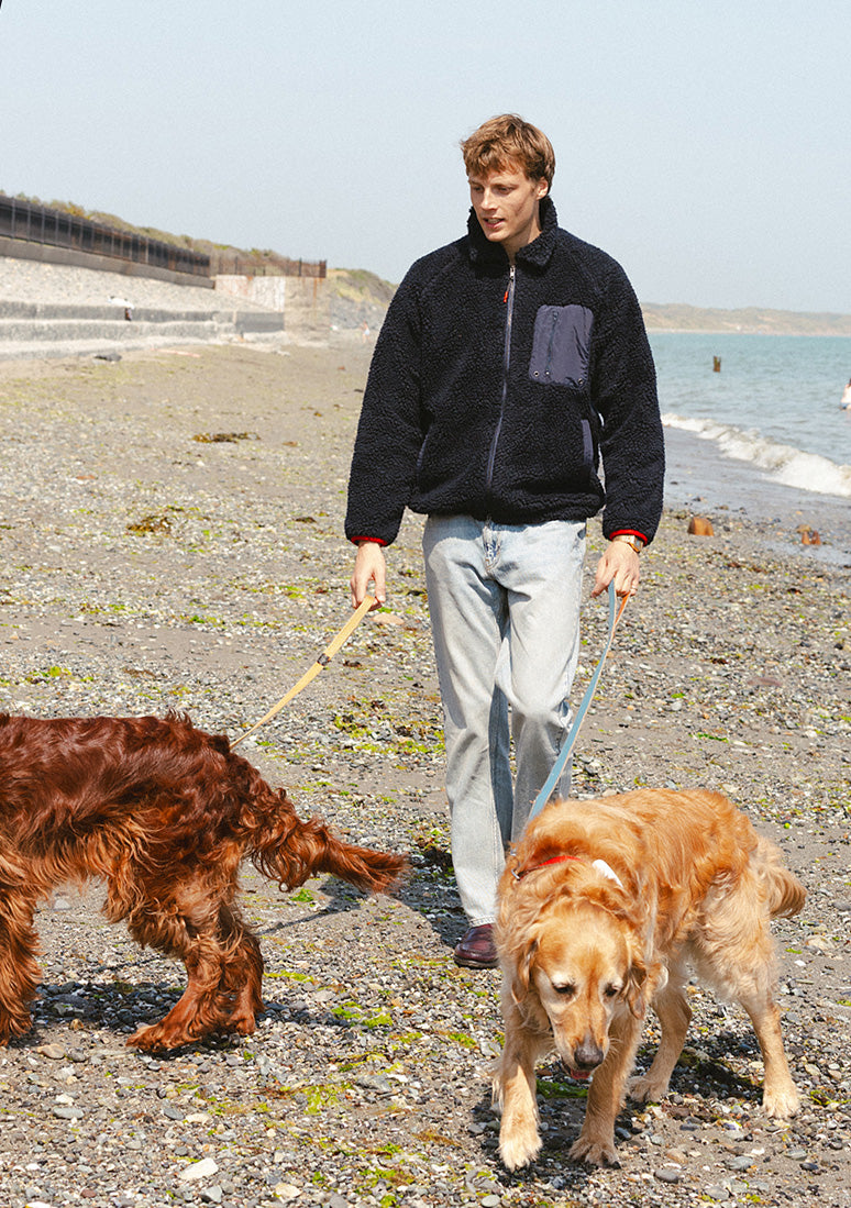 A model wearing a Percival navy wool blend fleece jacket and stone wash jeans while walking two dogs on a beach.