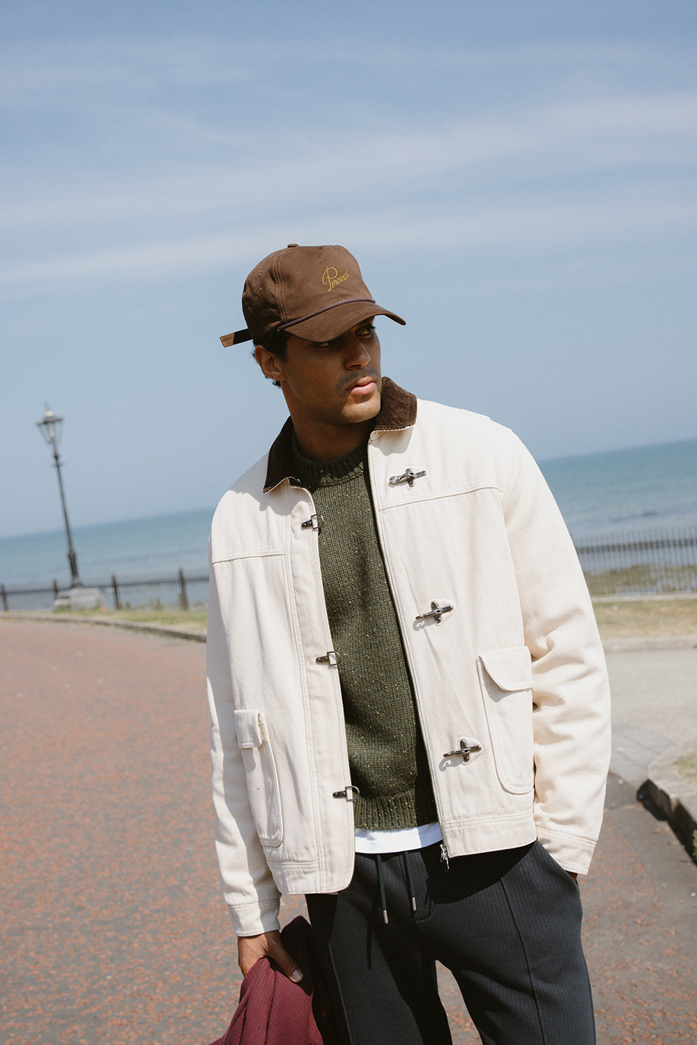 A model wears the cream Fontane Fireman Clasp Jacket by Percival with an espresso cap, dark knit jumper, and trousers.