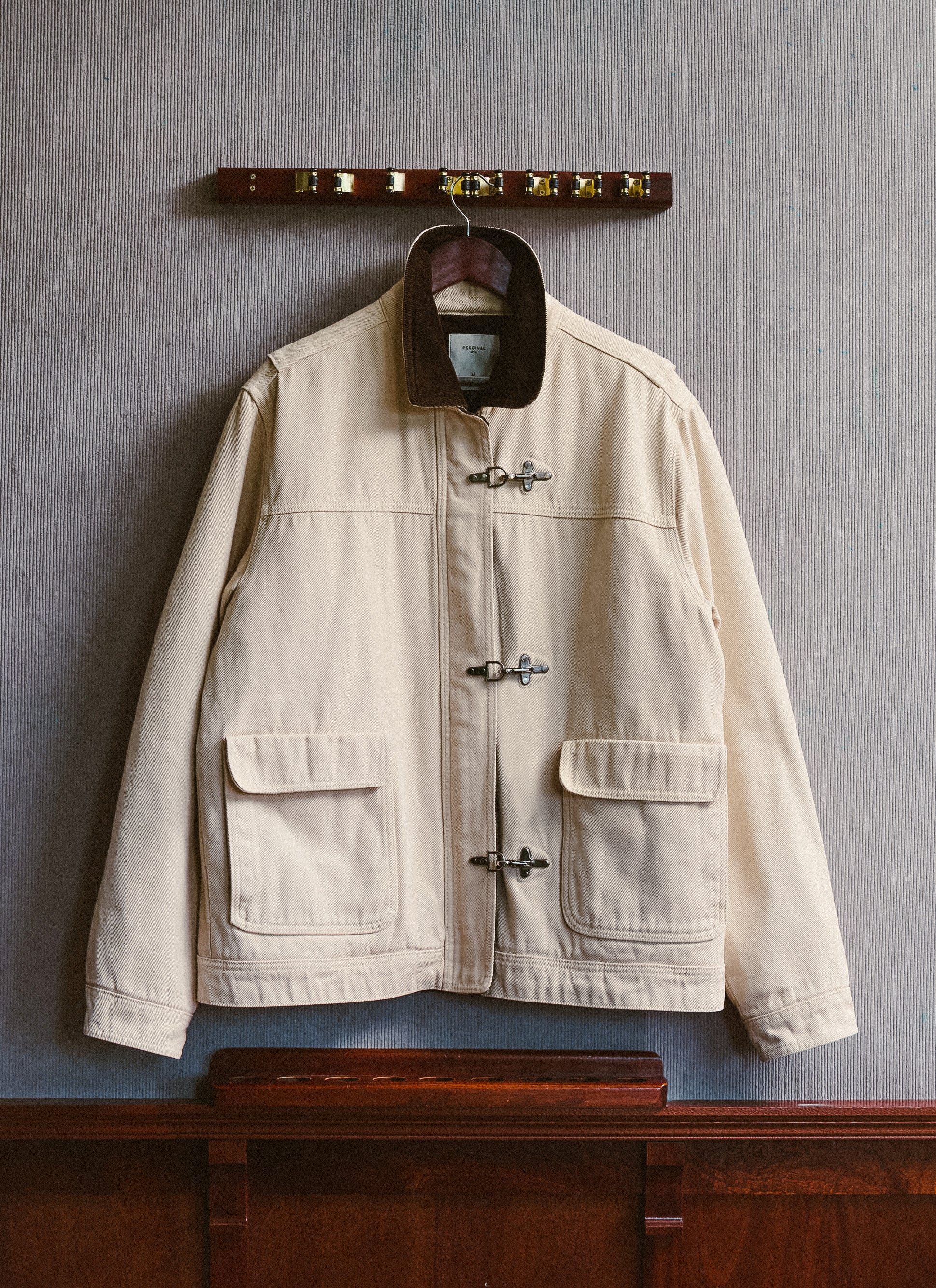 The cream Fontane fireman clasp jacket from Percival hanging on a wall, showing its corduroy collar and metal hardware.