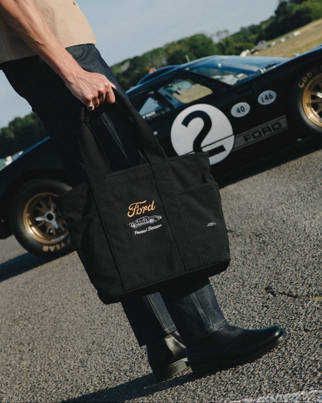 A person carrying a black Ford x Percival canvas tote bag on an outdoor racetrack, with a black race car blurred in the background.