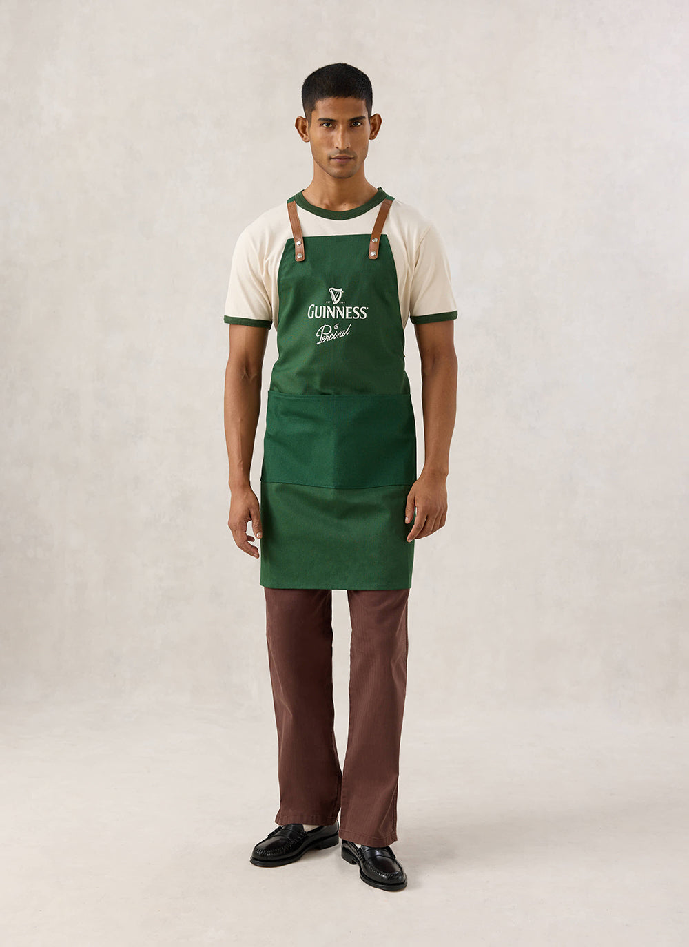 Model wearing the Guinness x Percival forest green apron with contrast tan straps and chest branding over a ringer t-shirt.