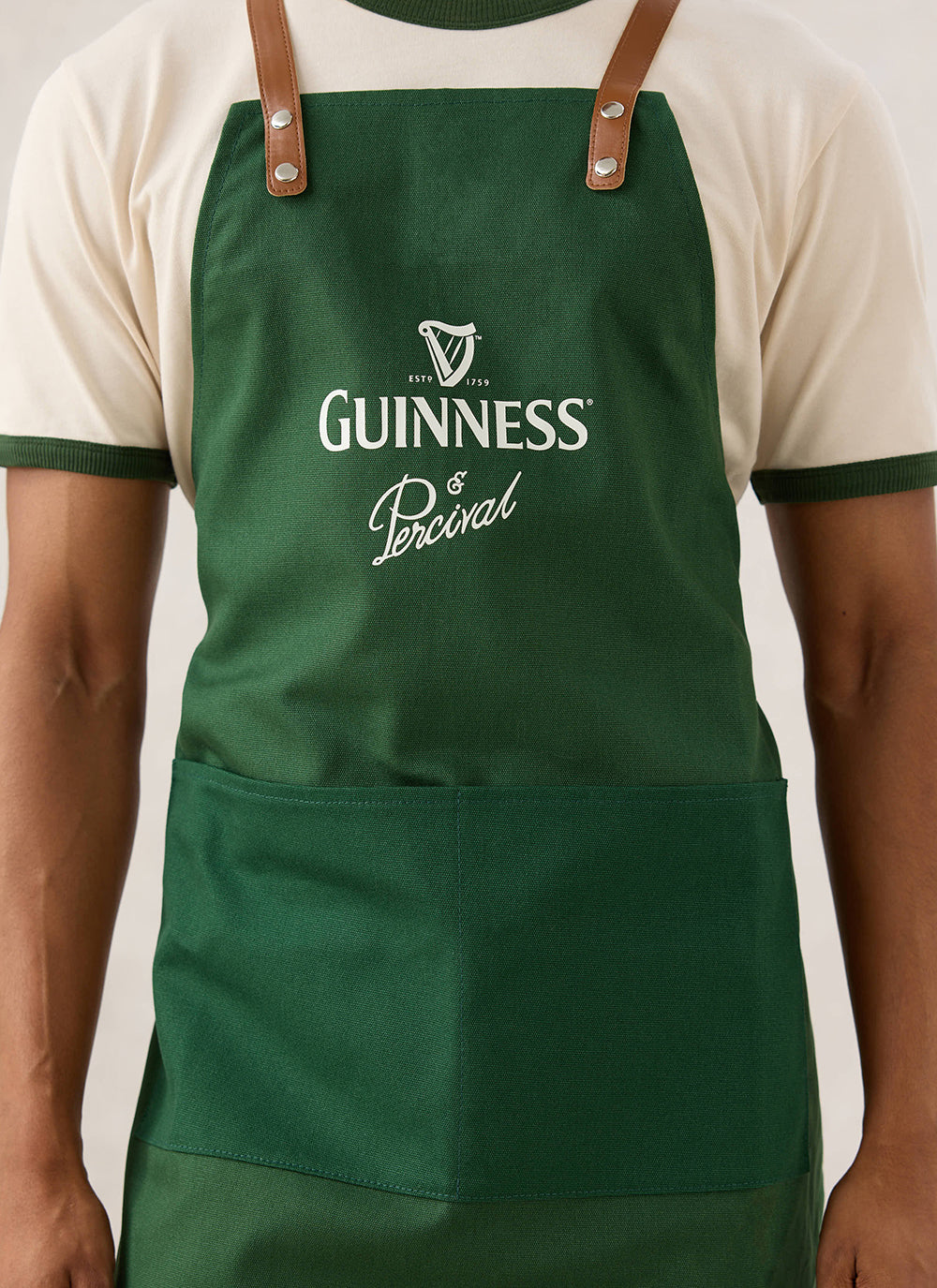 Close-up of a model wearing the forest green Guinness x Percival apron featuring contrast tan-look straps and chest branding.