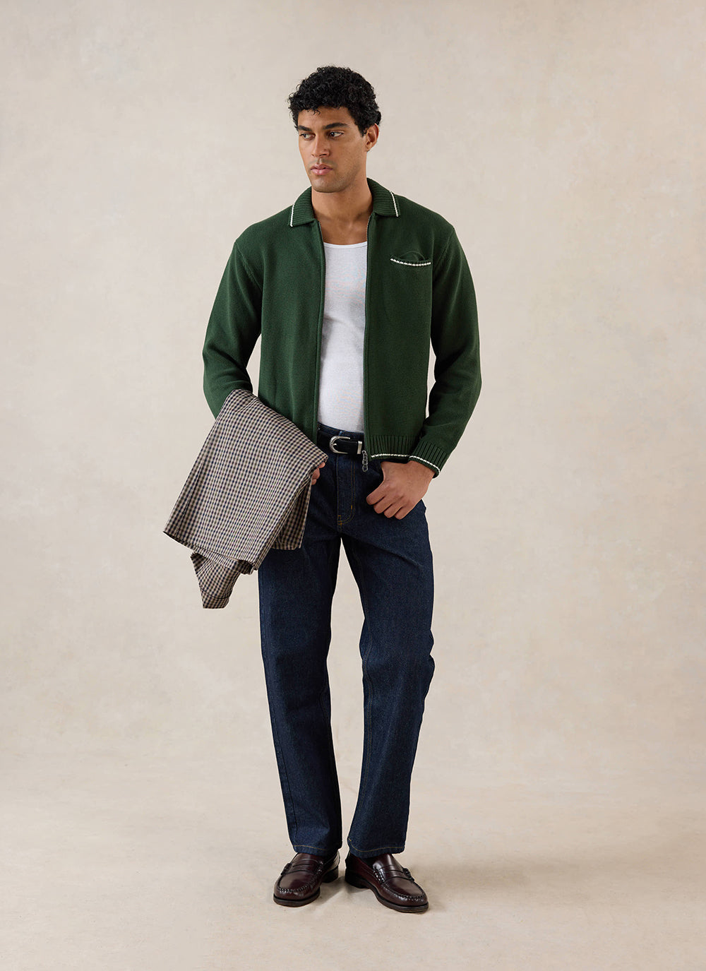 A model wears the Percival Gardner zip cardigan in forest green over a white vest and jeans, holding a checked jacket.