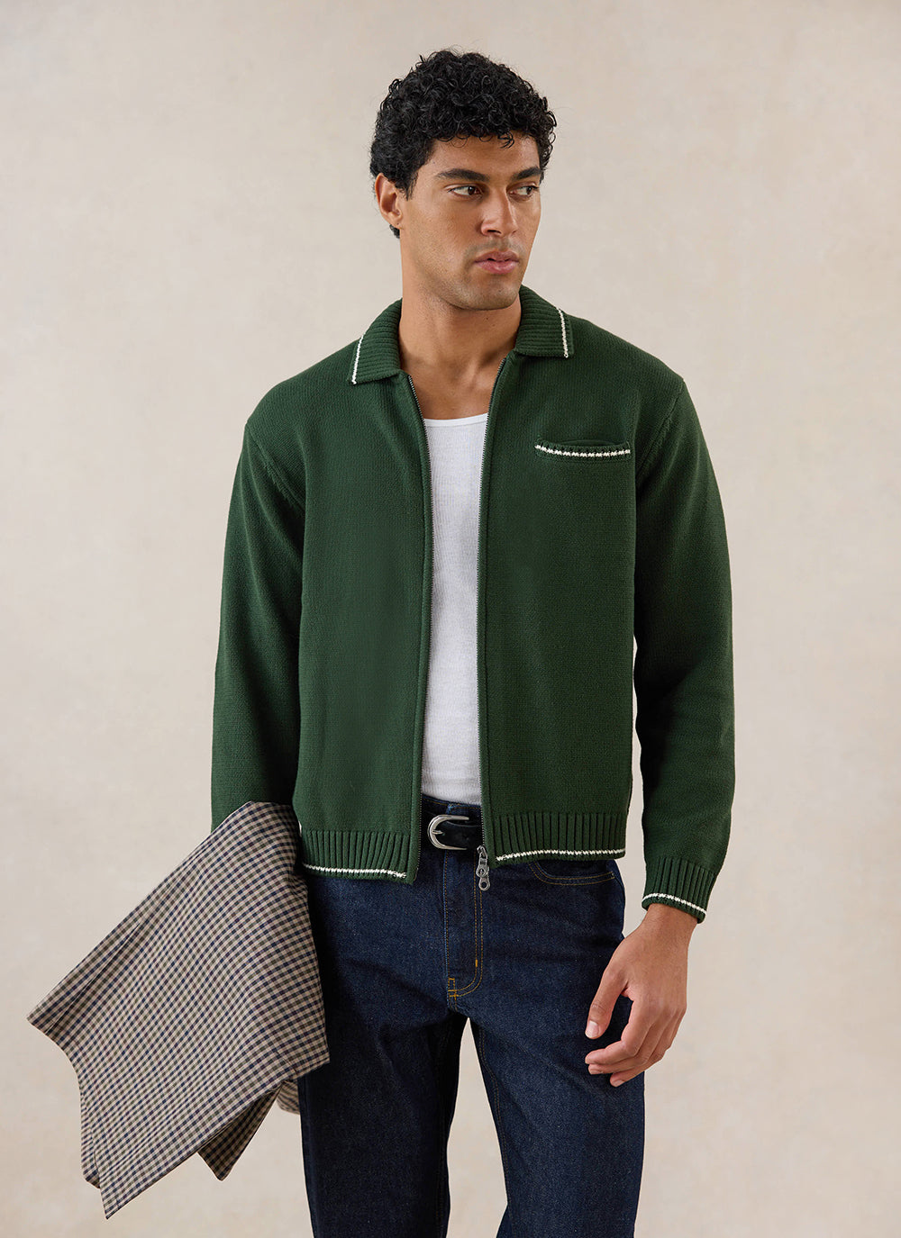 A model wears the forest green Percival Gardner knitted zip cardigan over a white vest, holding a checked jacket.