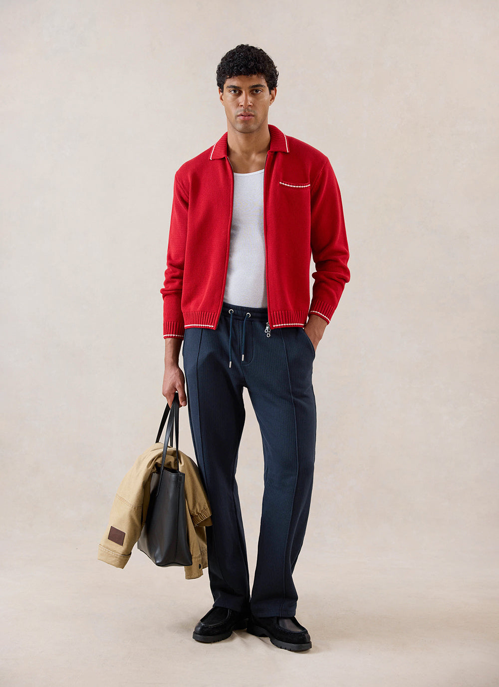 Model wearing the red Percival cotton knit zip cardigan with white vest and navy trousers, holding a black bag and jacket.