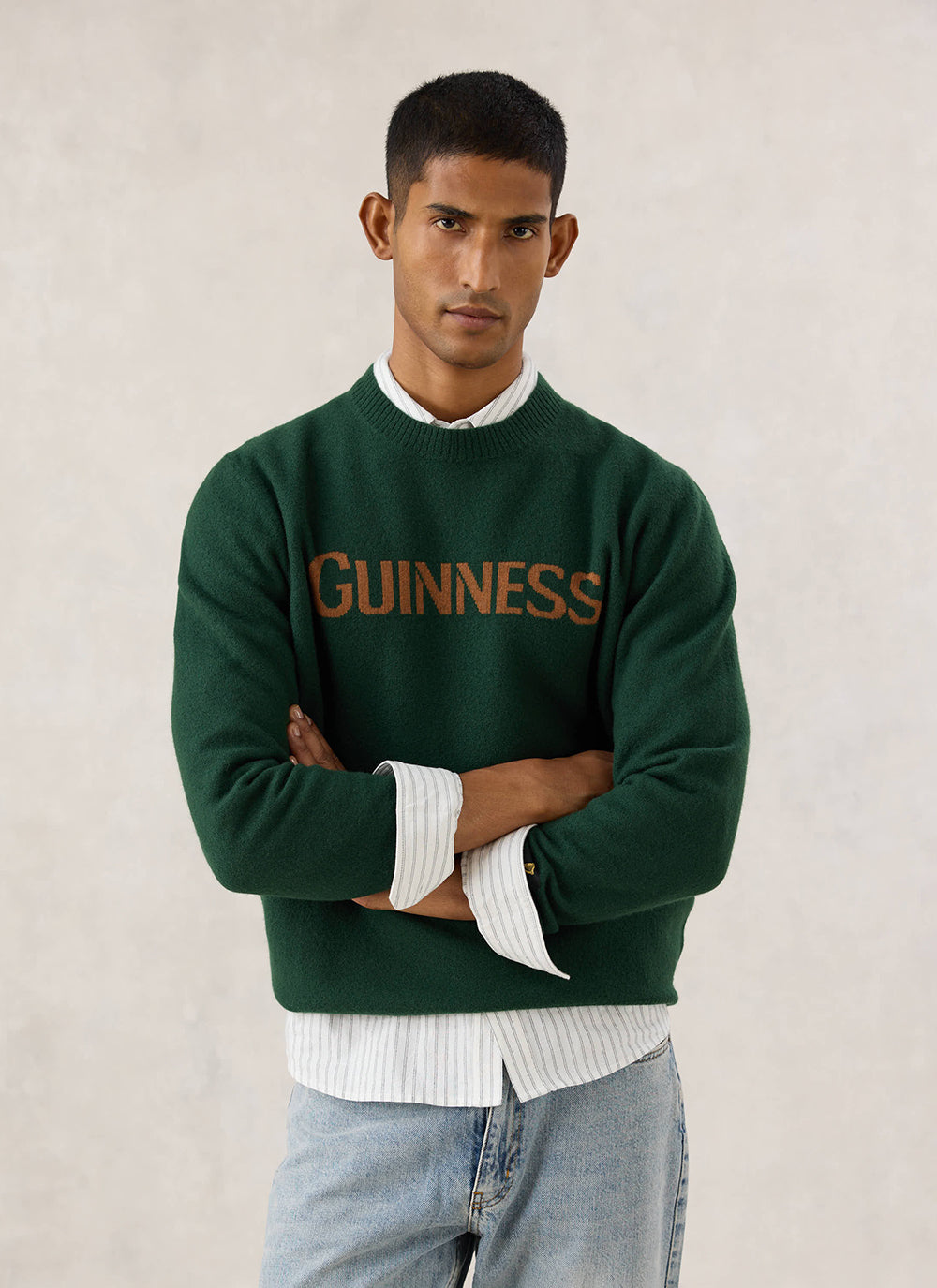 Model wearing the forest green Guinness x Percival merino wool jumper with jacquard branding over a striped shirt and jeans.