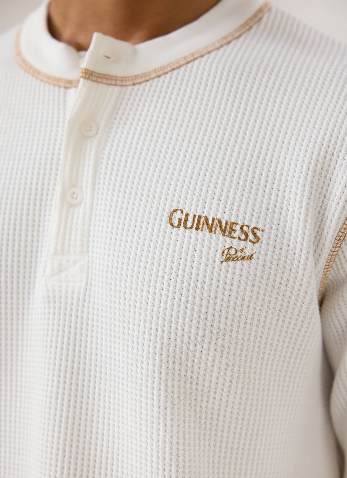 A close-up of the cream Guinness x Percival waffle henley, showing the button placket and printed chest logo.