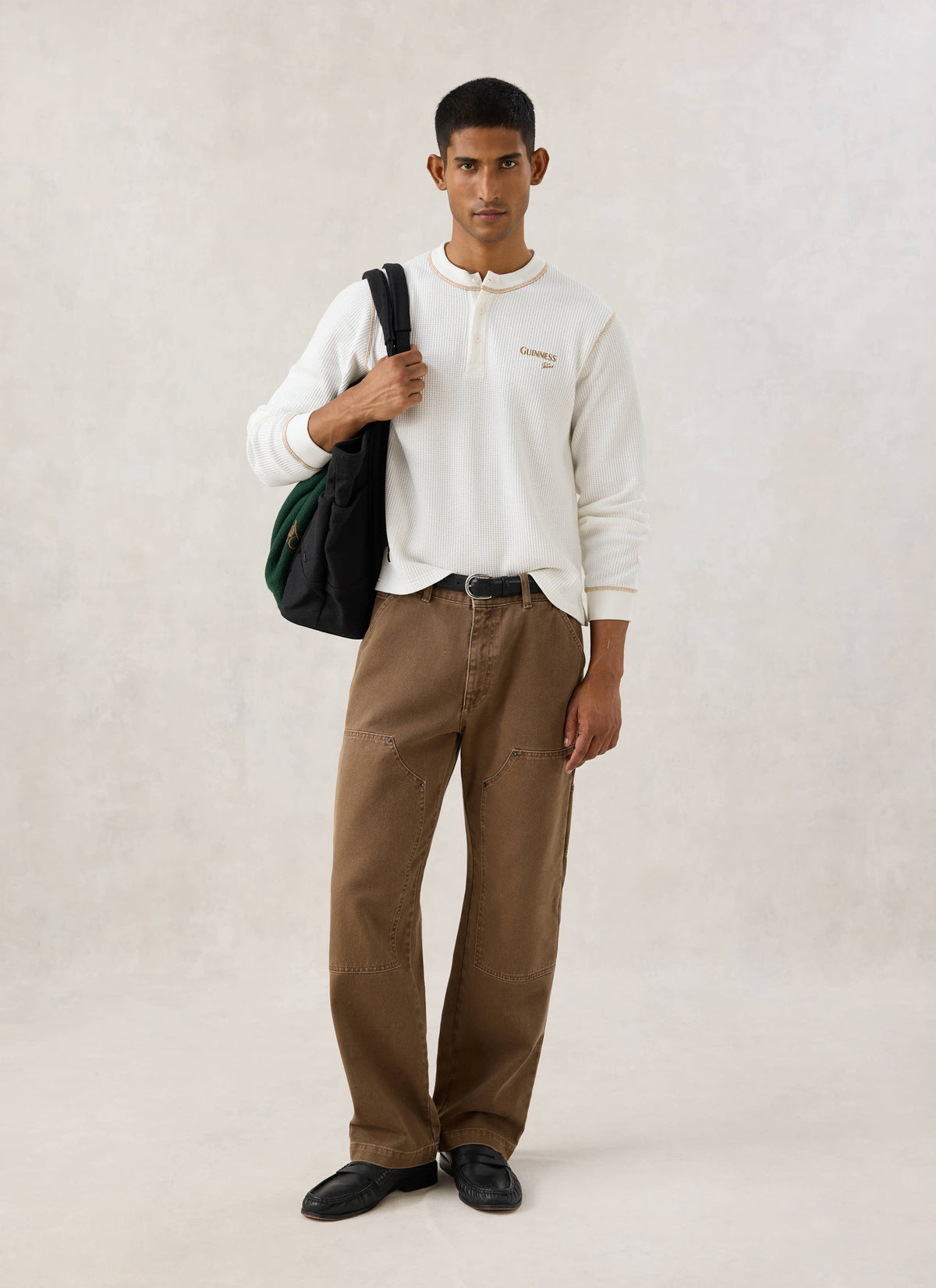 Model wearing the cream Guinness x Percival waffle henley long sleeve shirt with brown trousers and a black bag.