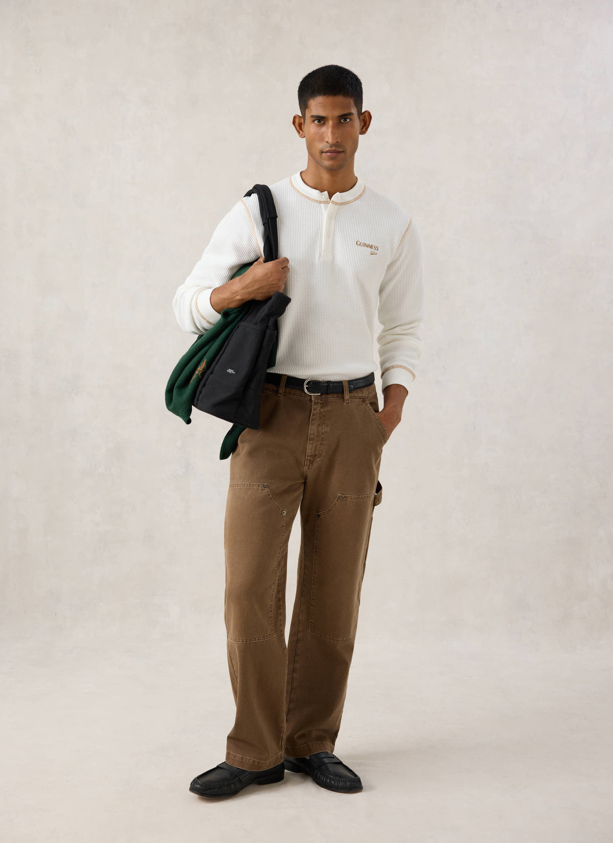 Model wearing the cream Guinness x Percival waffle henley with brown trousers and a black bag over his shoulder.