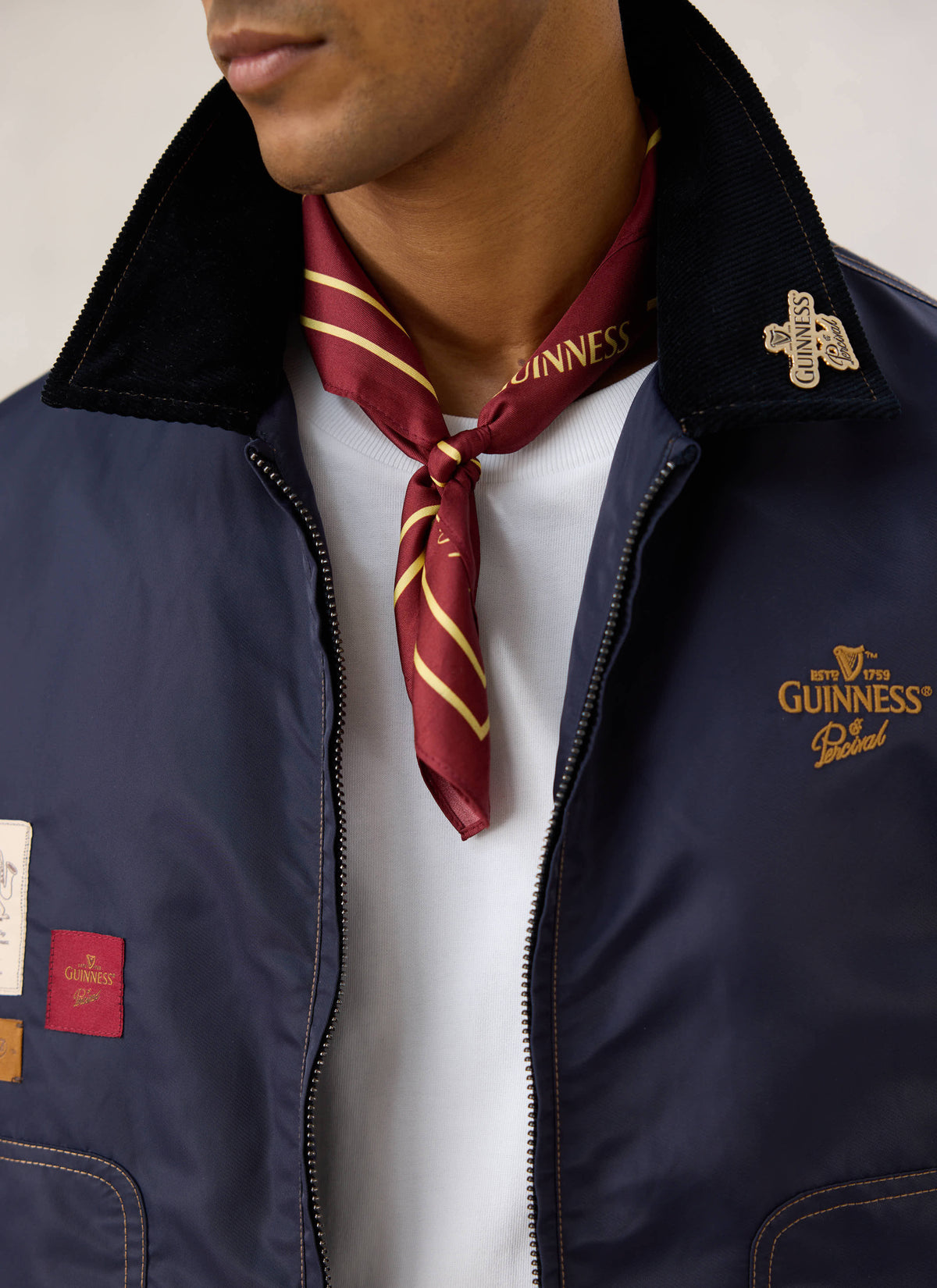 A model wearing the burgundy silk Guinness x Percival neckerchief tied at the neck under a navy jacket.
