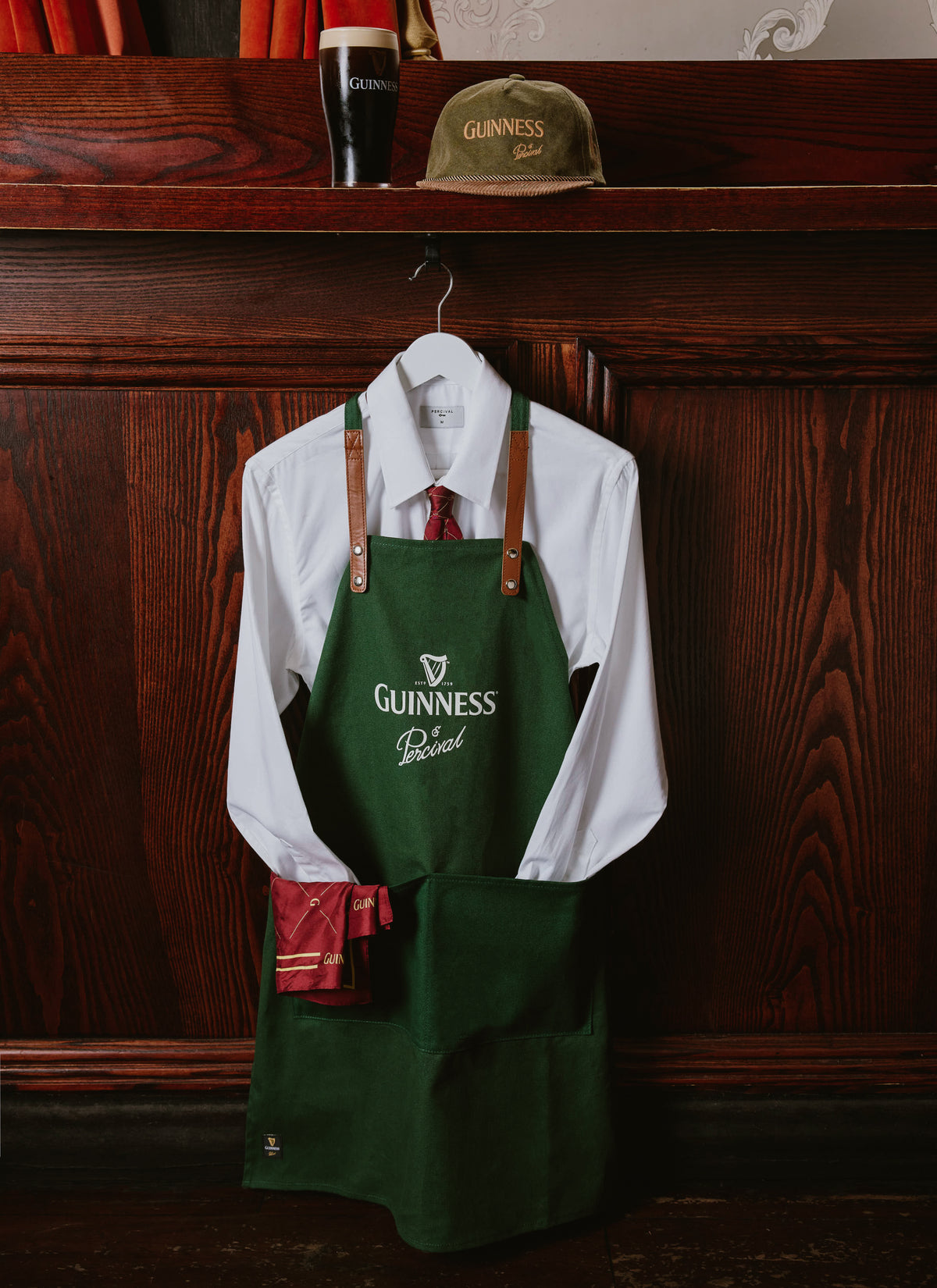 The Guinness x Percival forest green organic cotton apron hanging over a white shirt and tie against a wooden pub wall.