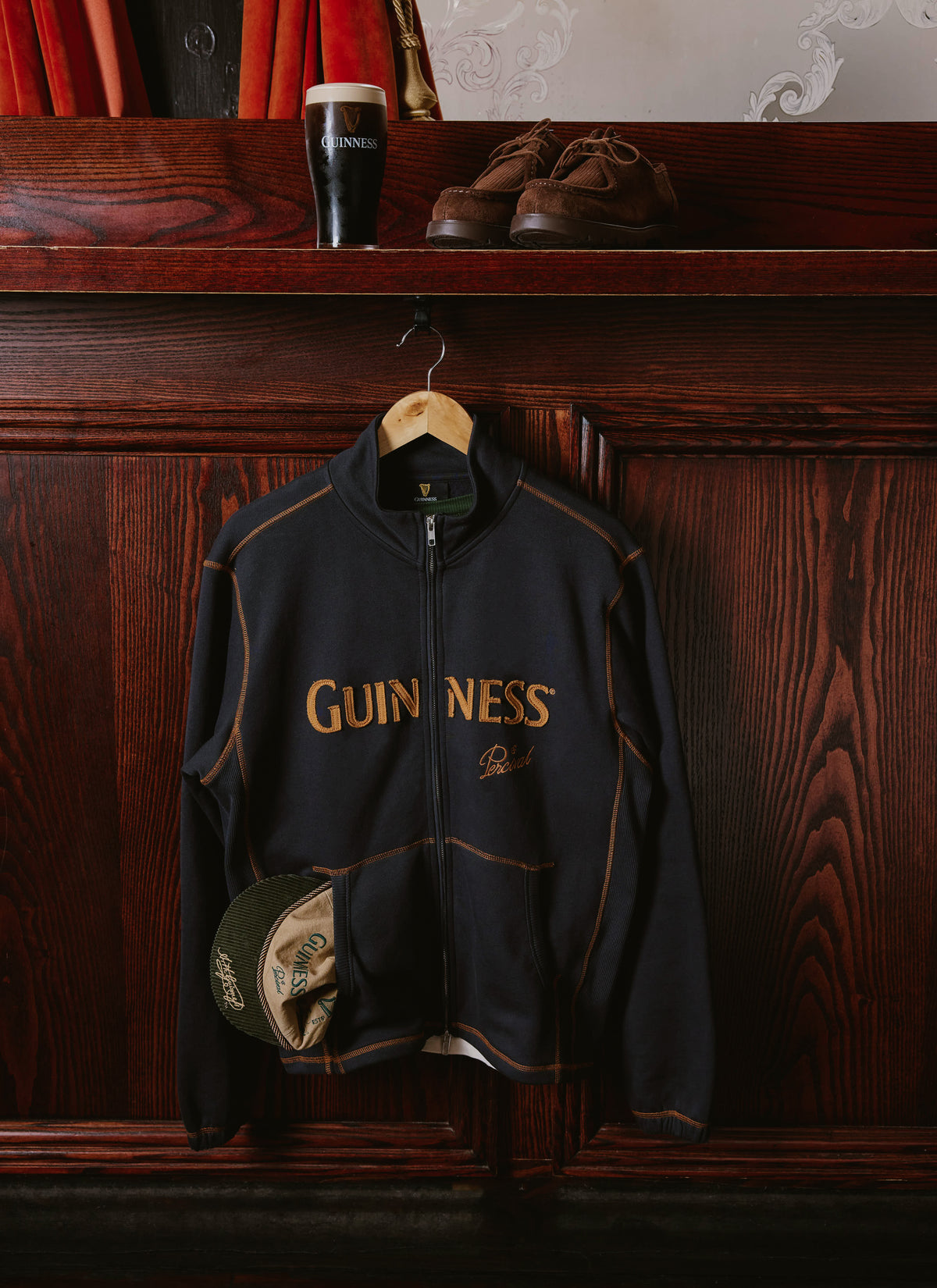 The navy blue Guinness x Percival Curtis funnel neck zip up sweatshirt hanging on a panel with contrast stitching.