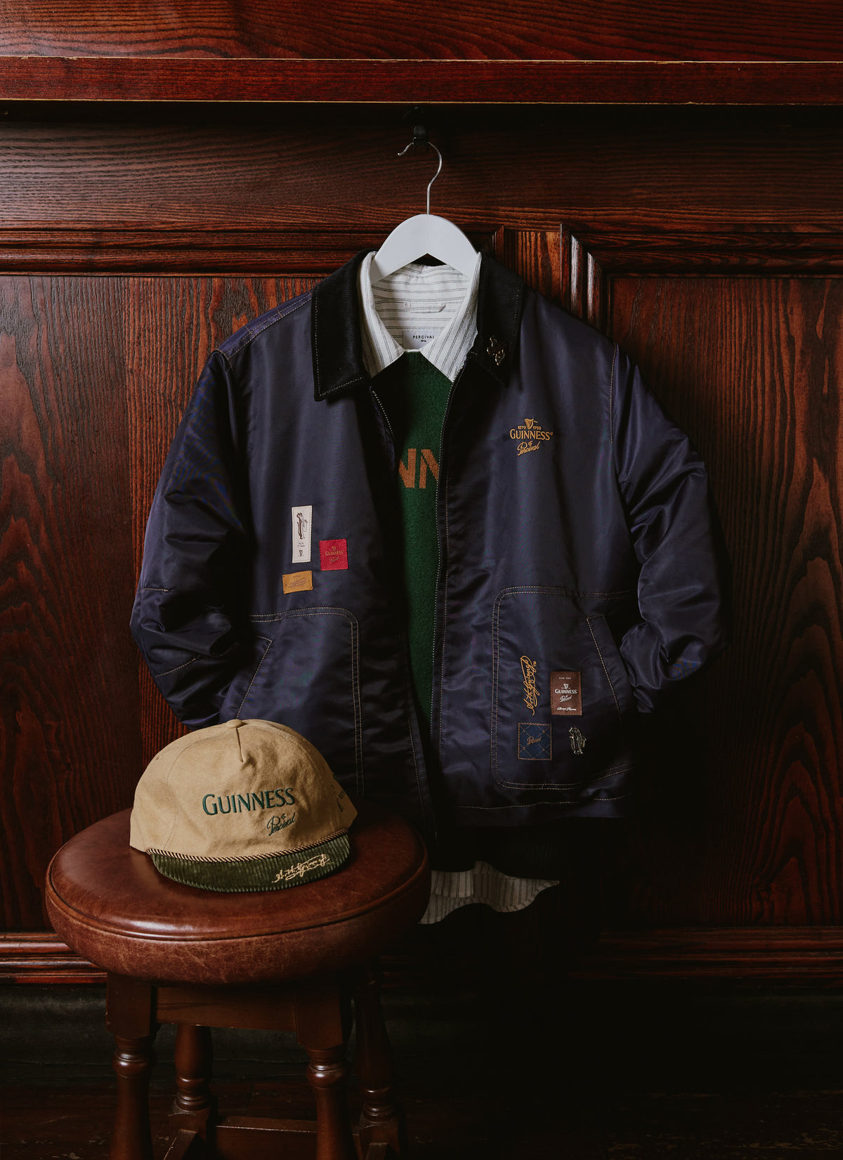 Guinness x Percival navy nylon bomber jacket and matching cap displayed against a dark wood pub interior.