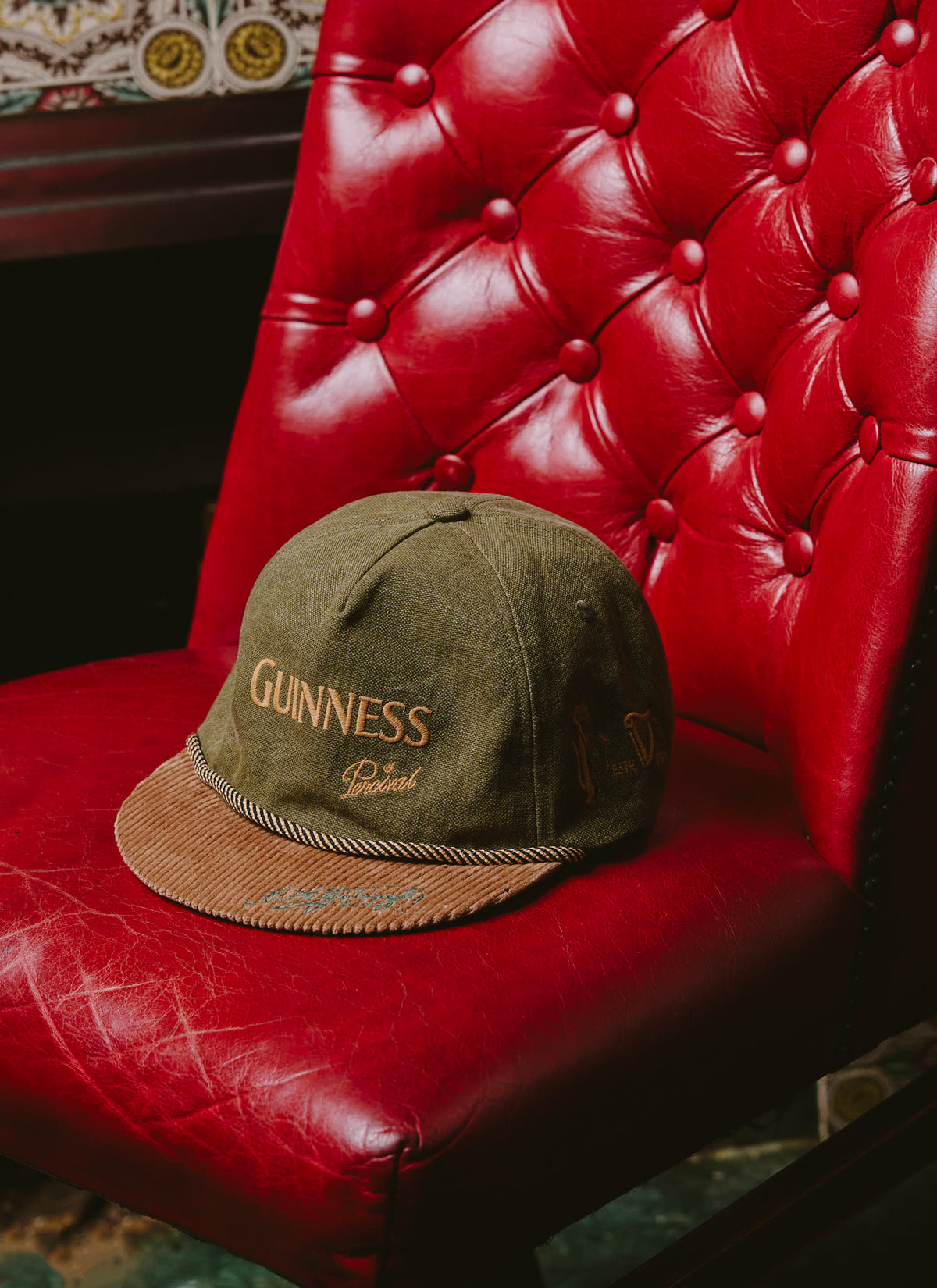 The Guinness x Percival green short peak cap with embroidered branding rests on a tufted red leather armchair.