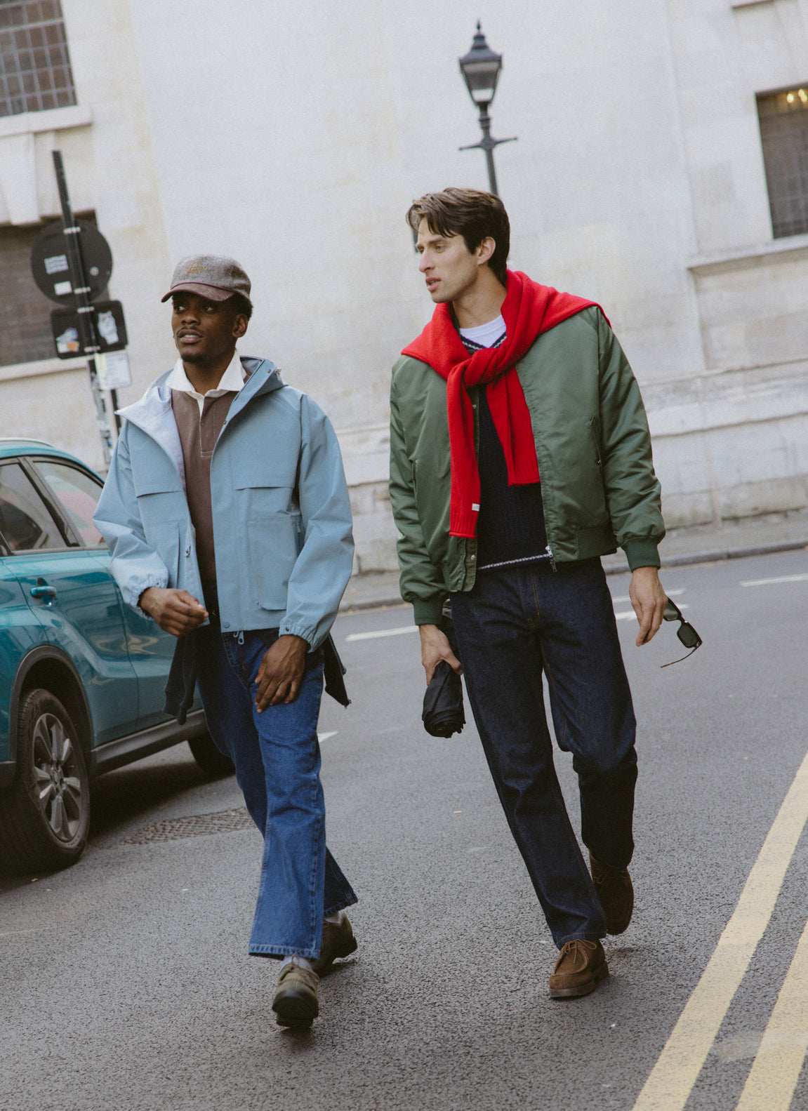 Two models walking outdoors on a city street, one wearing a green bomber jacket and a red jumper over the shoulders.
