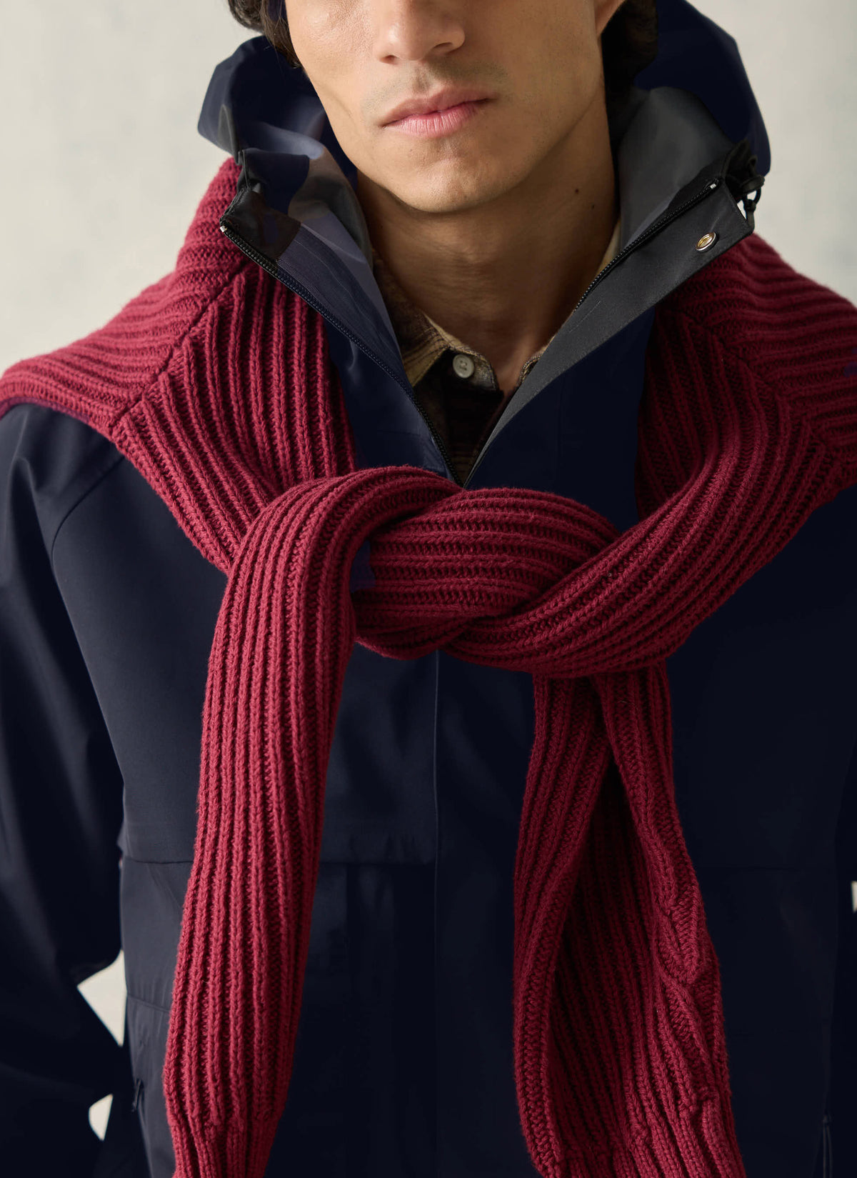 A close-up of a model wearing a burgundy ribbed knit sweater draped over the shoulders with a navy jacket and collared shirt.