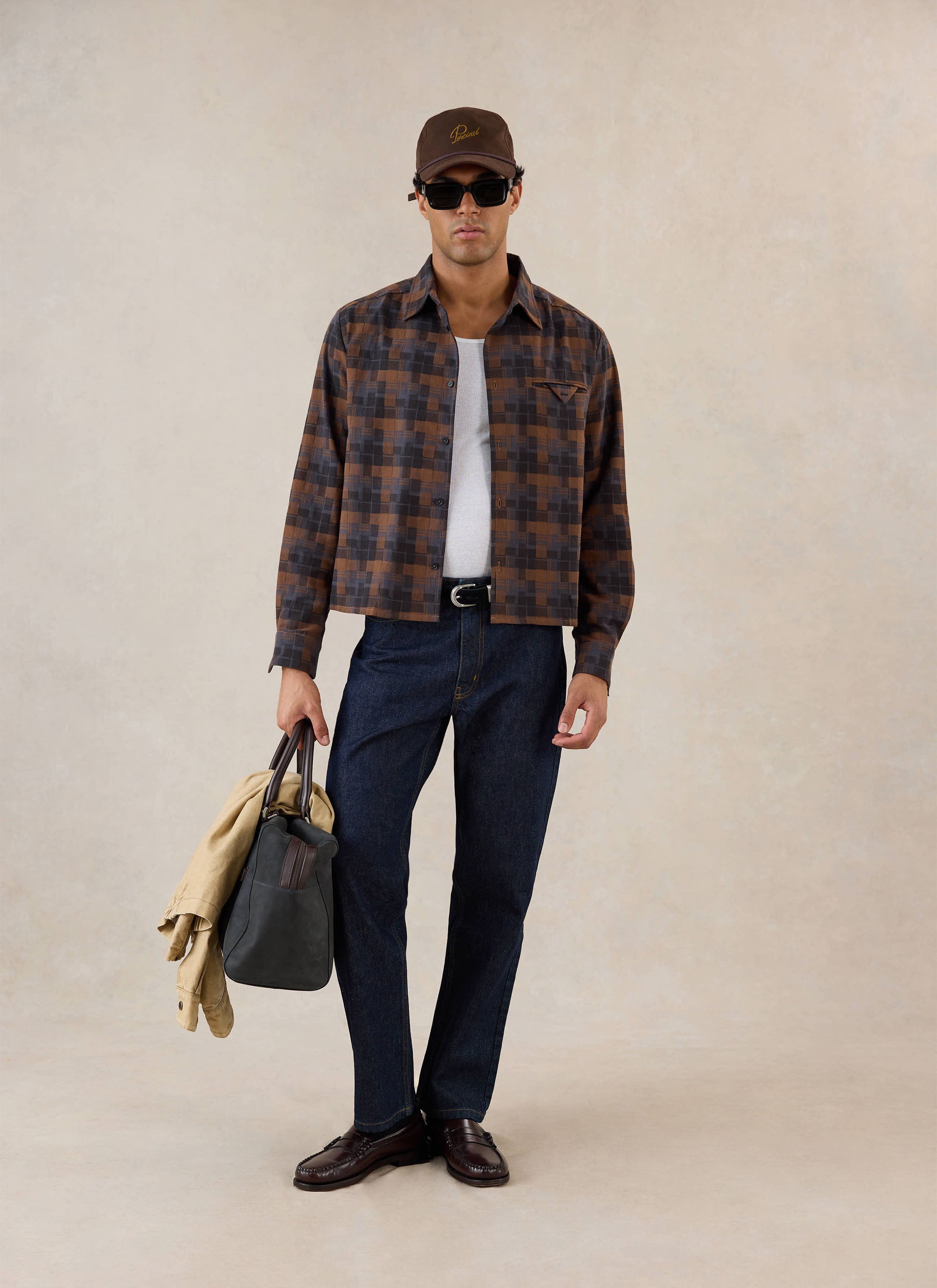 A model wears the brown check Percival shirt over a white vest, styled with dark jeans, sunglasses, cap, and a carry bag.