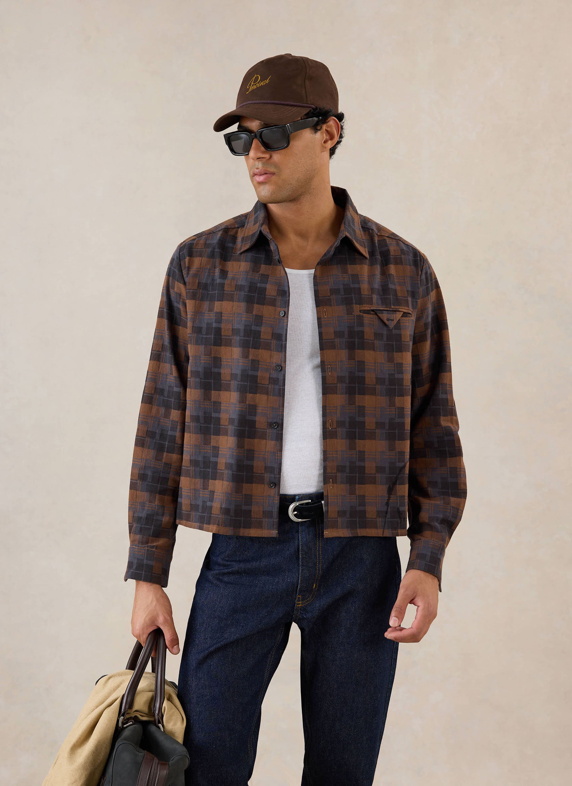A model wears the brown check Hinsley Boxy Long Sleeve Shirt from Percival over a white vest with sunglasses and a brown cap.